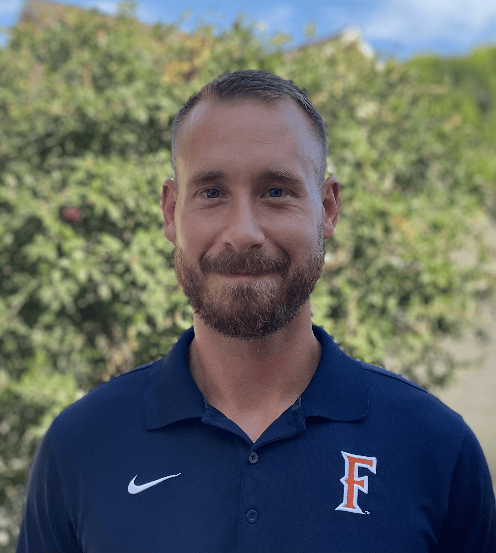Close-up of a smiling man with a beard and short hair, wearing a dark blue sports polo shirt with a logo that includes a large letter 'F' and a Nike swoosh, standing outdoors with trees and a blue sky in the background.
