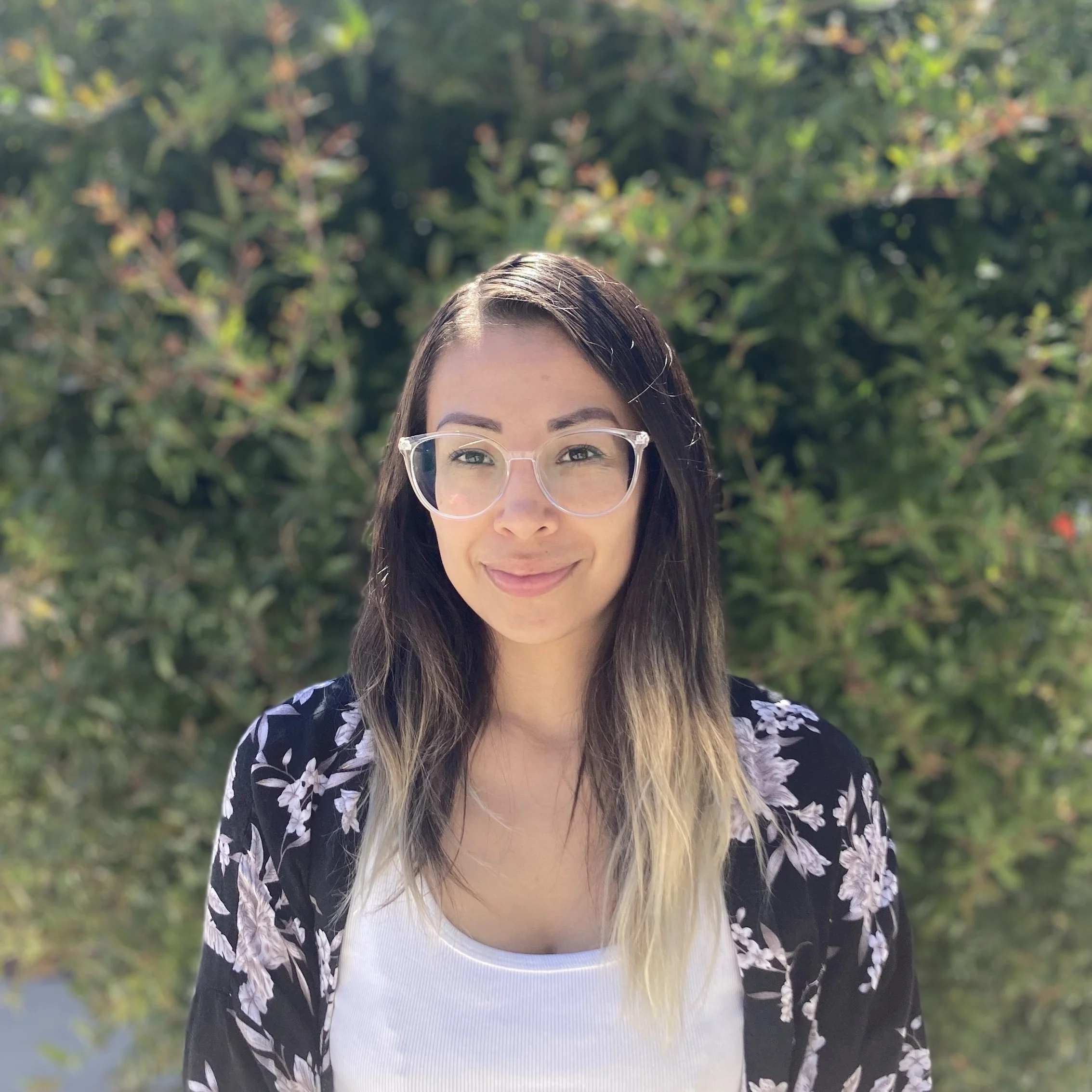 A woman with long hair wearing clear glasses, a white shirt, and a black floral jacket, standing outdoors in front of green foliage.