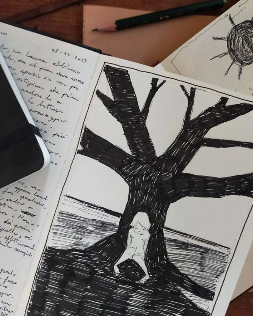 A black ink drawing of a large tree with a girl sitting at its base near the water, on a white sheet of paper.