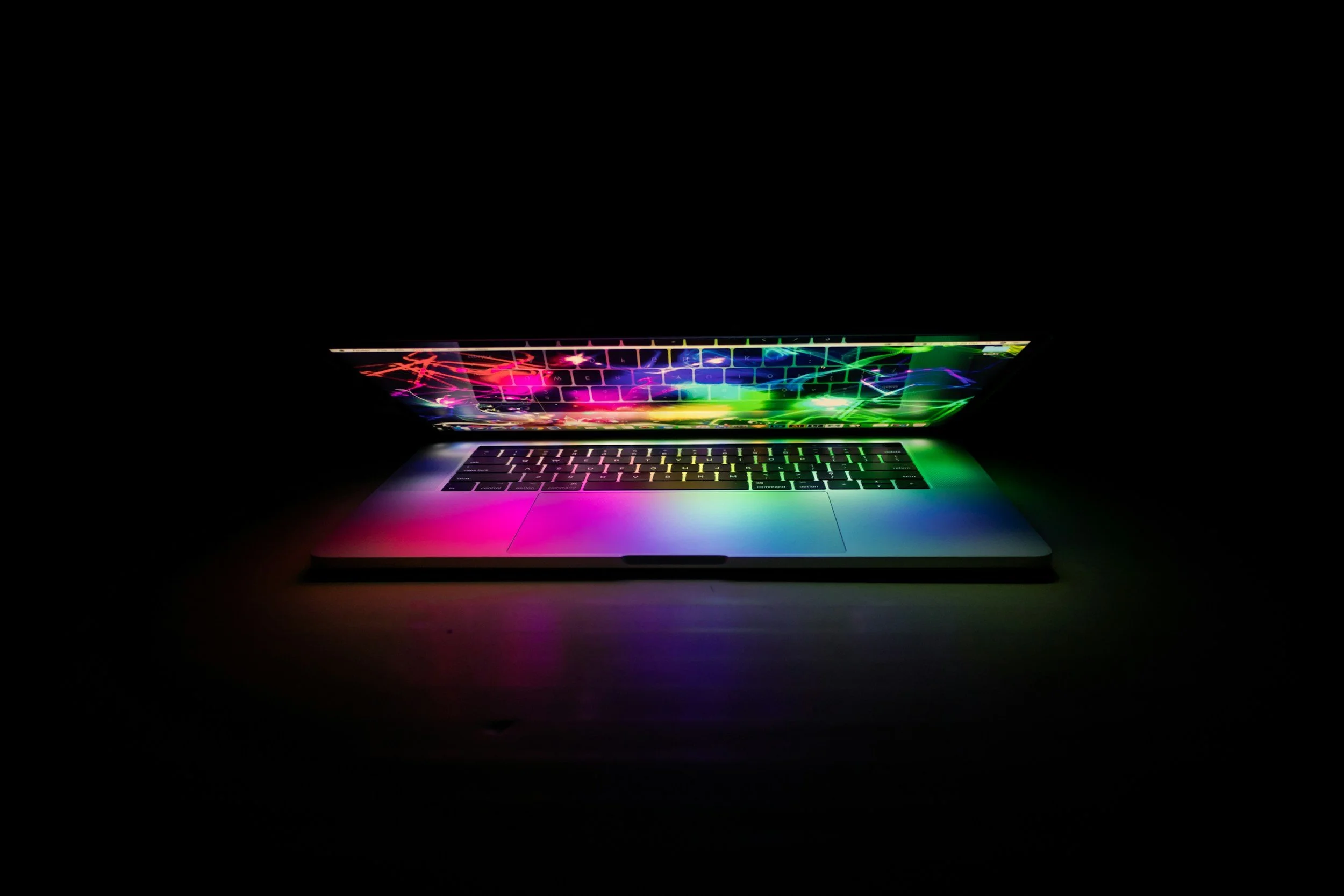 A laptop with a rainbow-colored backlit keyboard in a dark room, displaying a colorful, glowing screen.