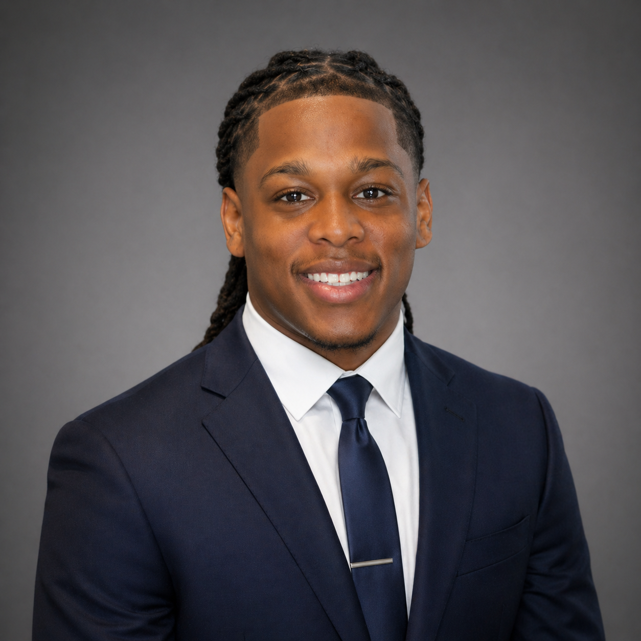 Young man in navy suit, white shirt, and navy tie smiling in a professional headshot against a gray background.