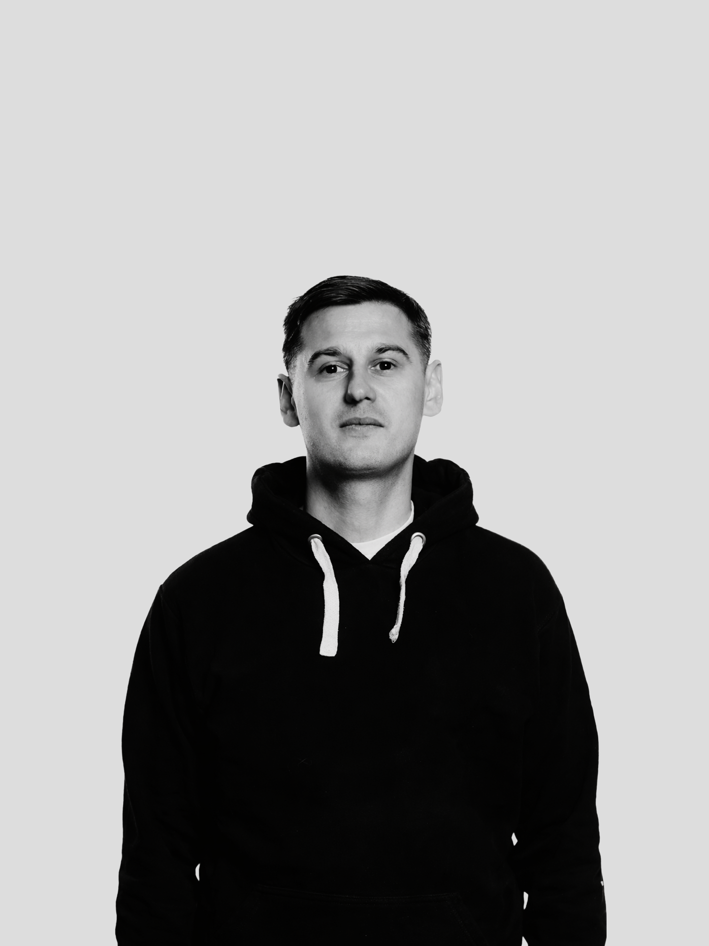 A black and white photo of a young man with short dark hair, wearing a black hoodie with white drawstrings, standing against a plain light gray background.