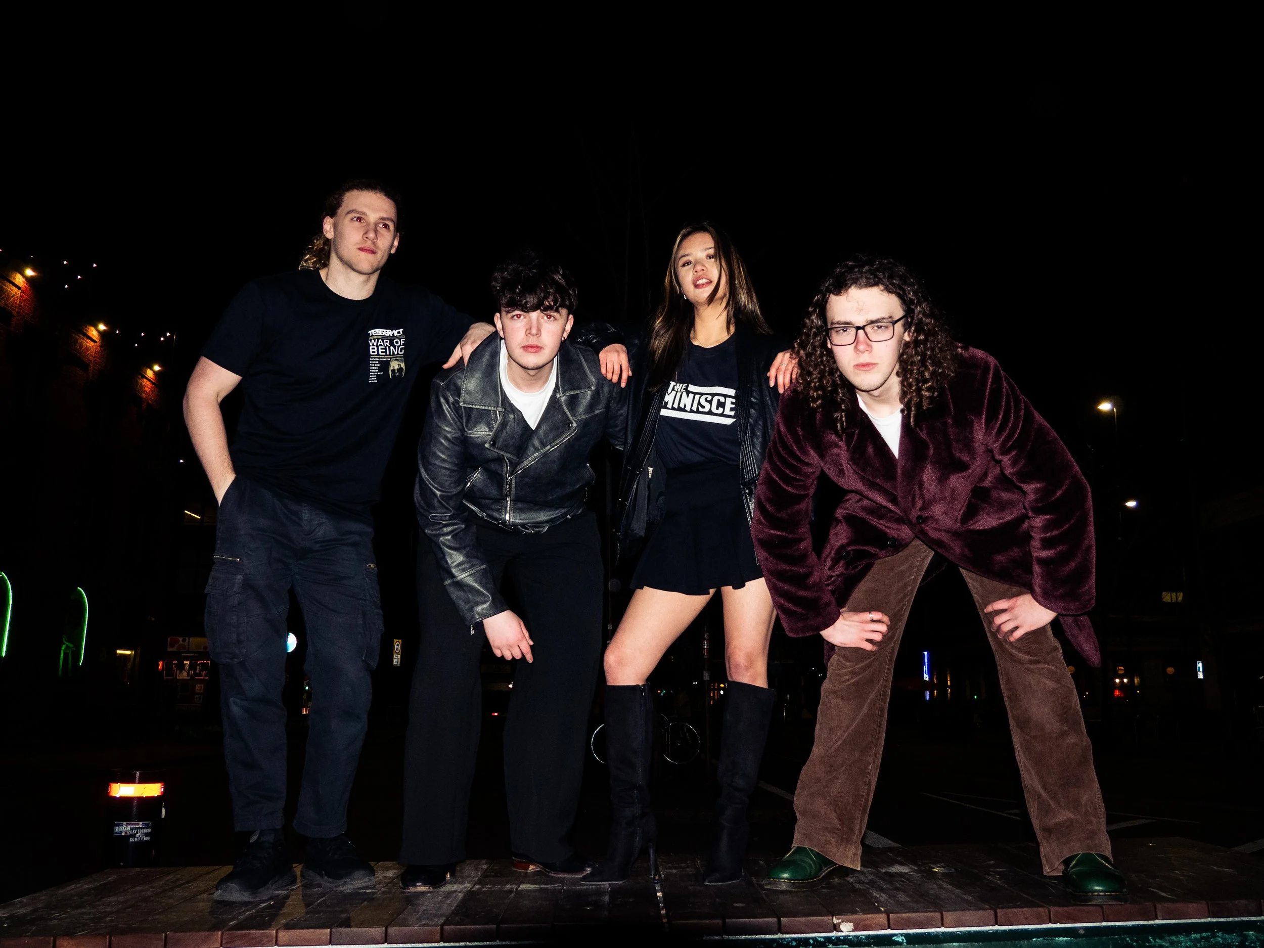 Four young adults, two men and two women, posing together outdoors at night under a dark sky with a few streetlights in the background.