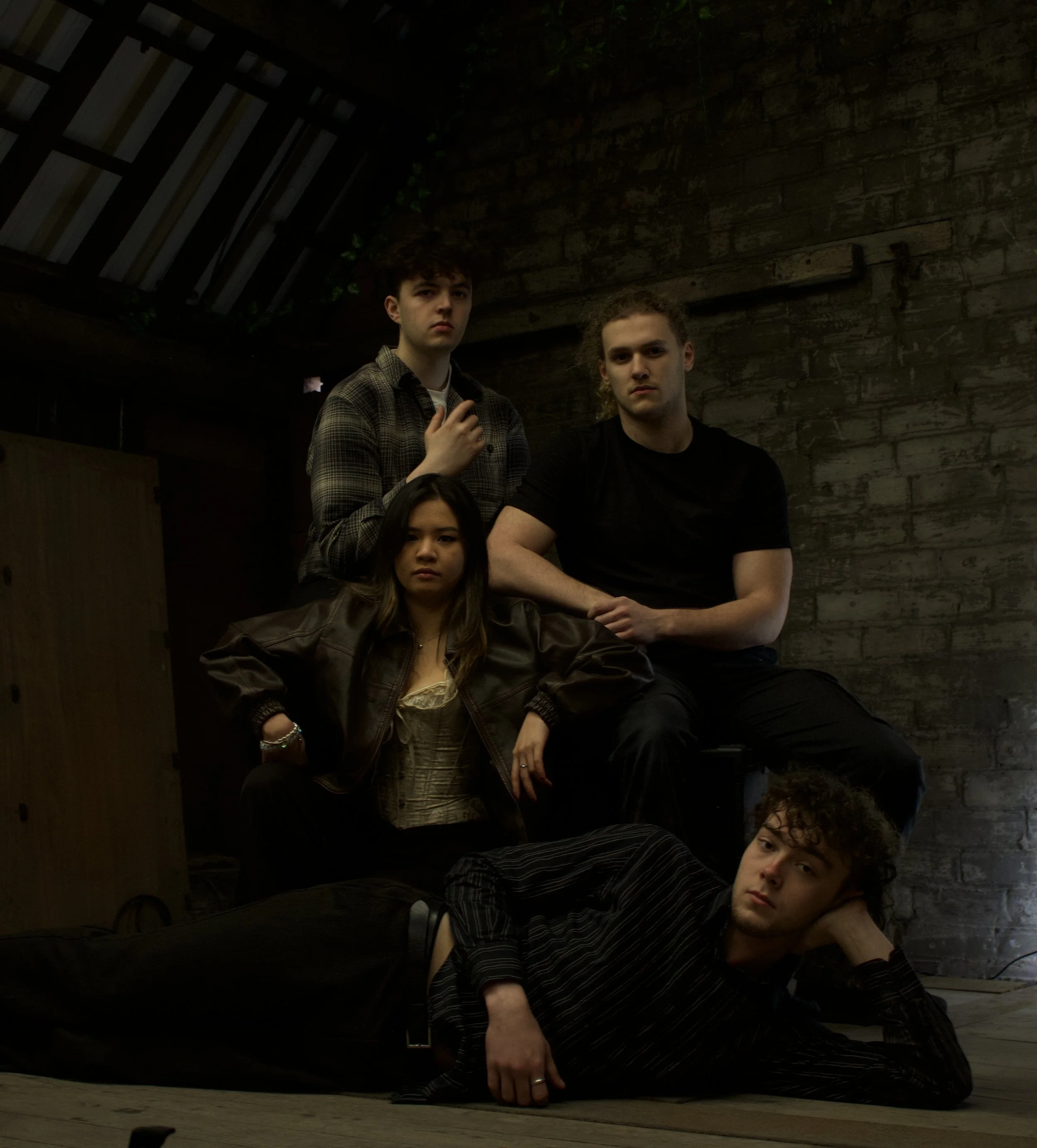 Four young people posing indoors against a dark brick wall with wooden stairs in the background. The group includes two males and two females, dressed casually.