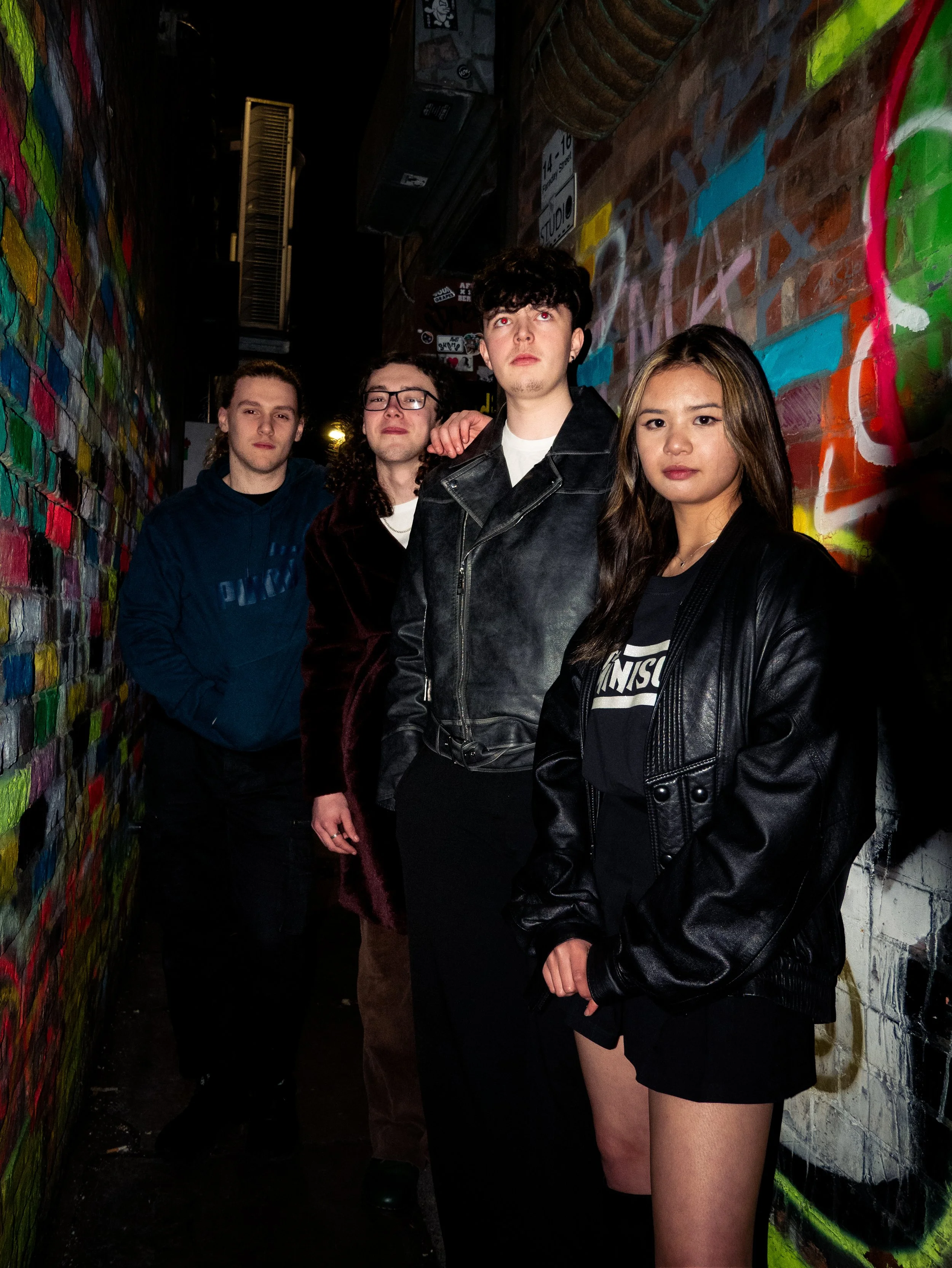 Four young people stand in a dimly lit alleyway with colorful graffiti on the walls, posing for a photo. The person in front is a young woman with long hair, wearing a black leather jacket and a black oversized T-shirt. Behind her, three young men are visible, one wearing glasses and a burgundy jacket, another with curly hair and a white shirt, and the third with short hair and a dark sweater. The scene has a gritty, urban vibe.