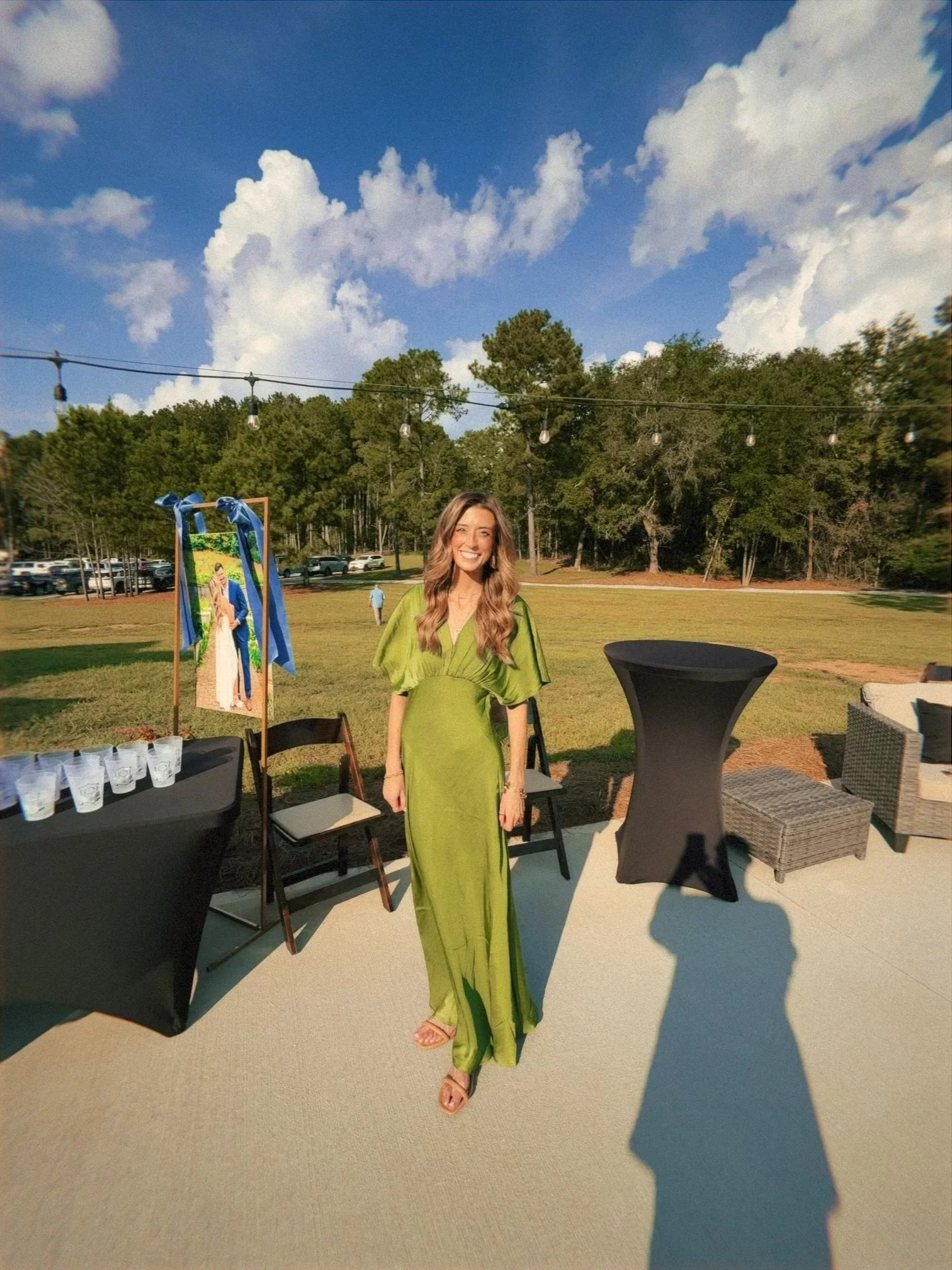 A woman with long wavy hair wearing a bright green dress, smiling, standing outdoors at an event with tables, chairs, and a string of lights in the background, under partly cloudy blue sky.