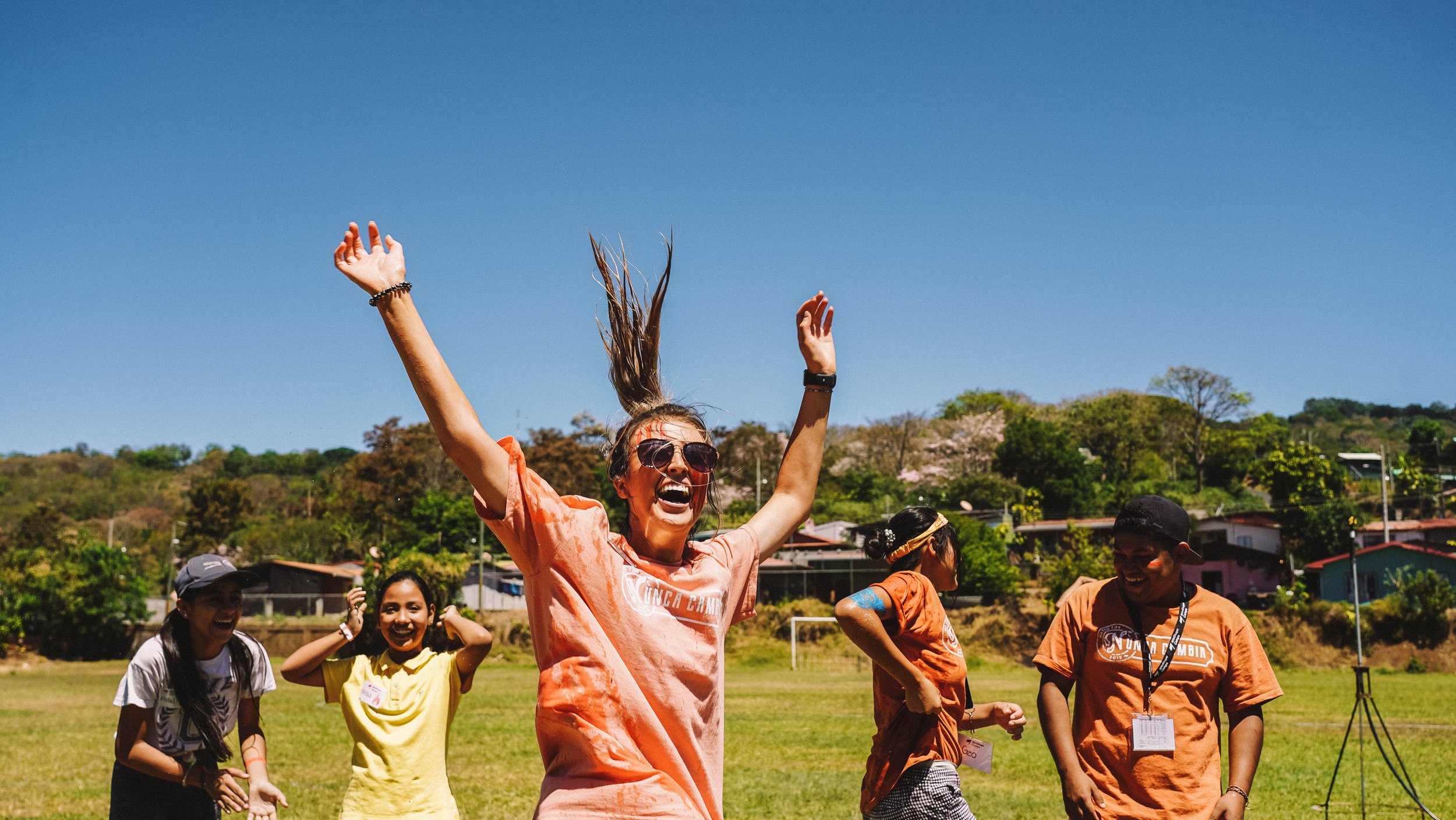 A group of smiling people are outdoors on a grassy field, celebrating or having fun, with clear blue sky and trees in the background. One woman with sunglasses and an orange shirt is raising her arms joyfully.