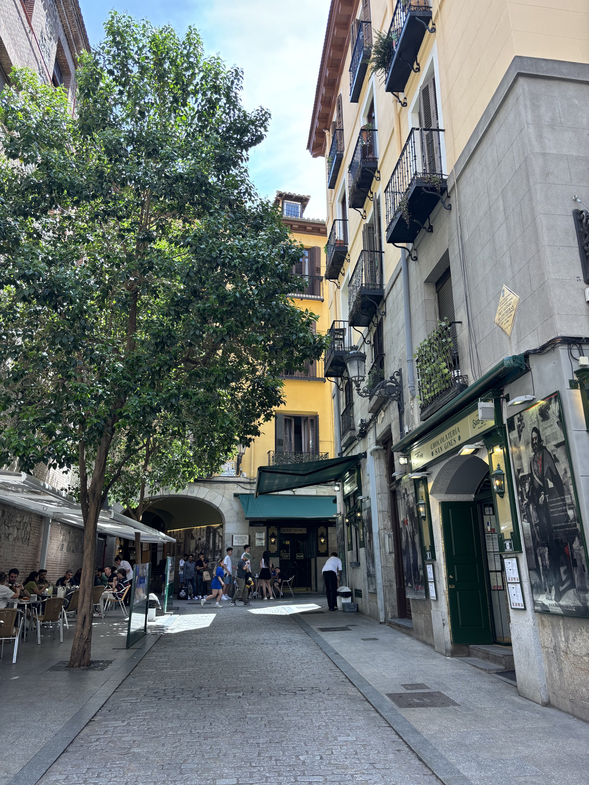 A lively outdoor scene in a European city, with people dining at tables on a street lined with colorful buildings, trees, and a cafe entrance.