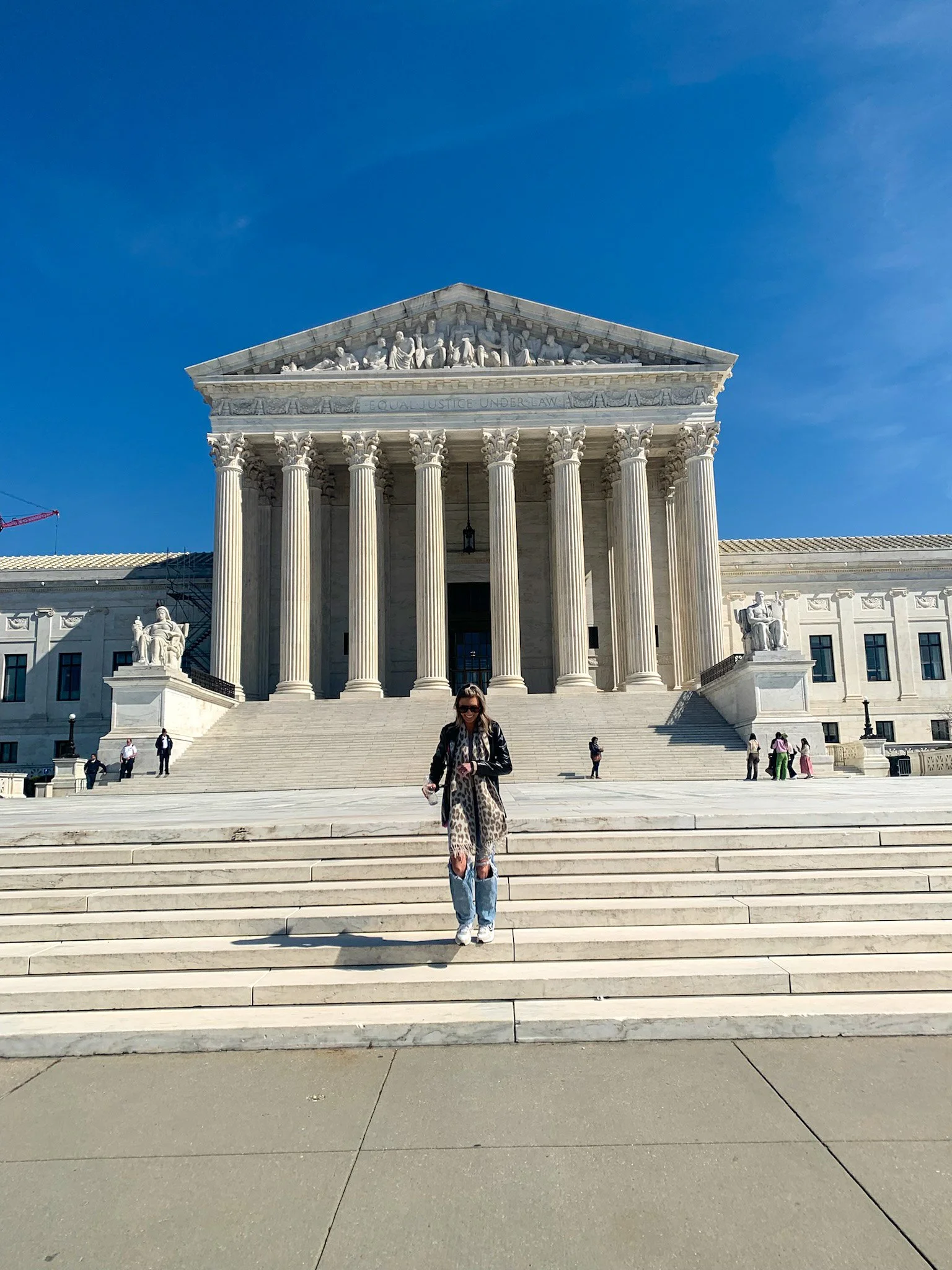 A woman standing on the steps in front of the United States Supreme Court building in Washington, D.C., with the pediment and columns visible behind her, under a clear blue sky.