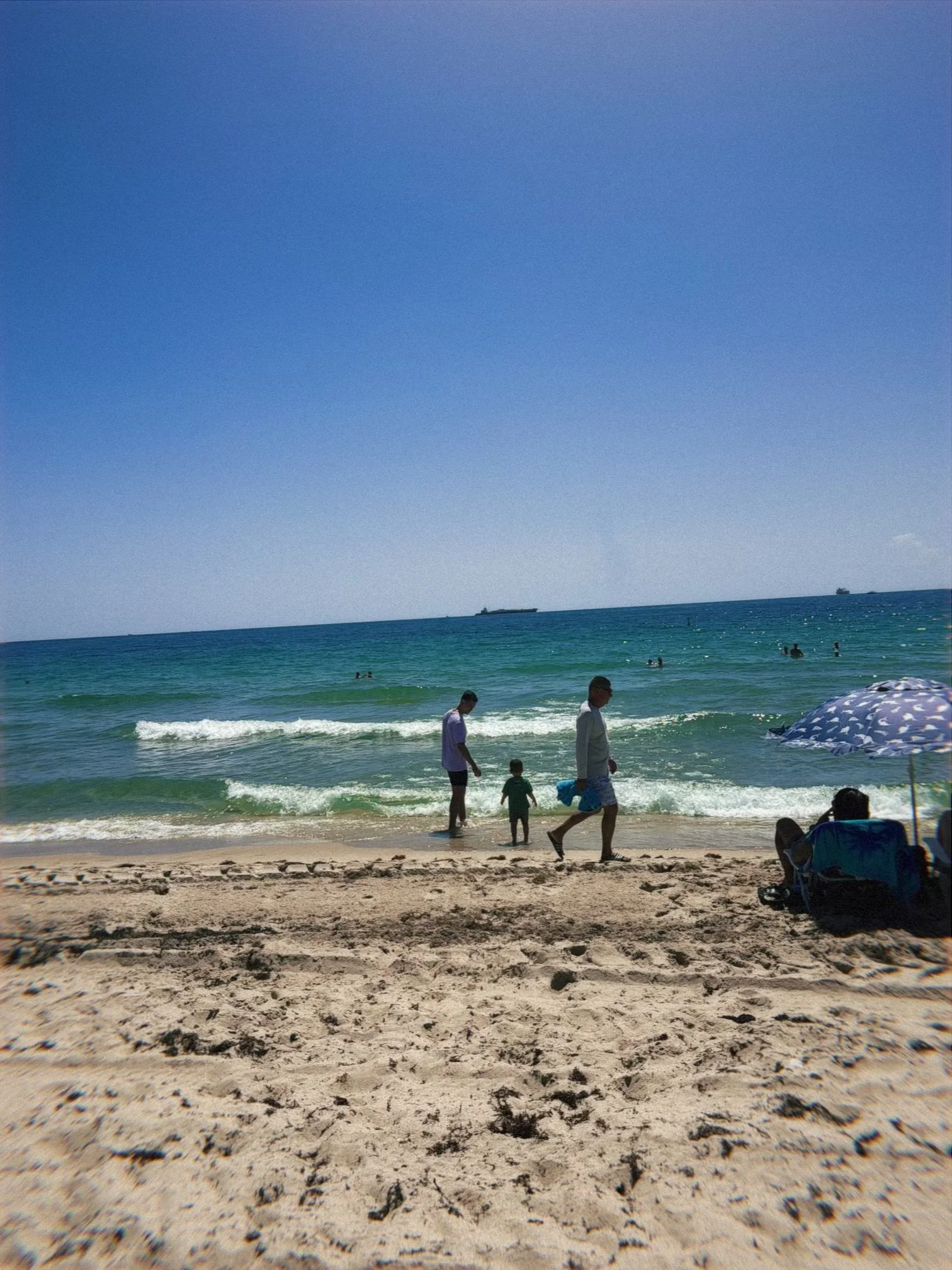 People enjoying a sunny day at the beach, walking along the sandy shore near the ocean with a clear blue sky overhead. Some are swimming in the water, while others relax under umbrellas.