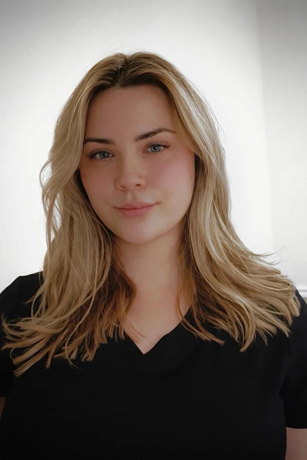 A young woman with blonde wavy hair, wearing a black top, standing in front of a plain white wall, looking at the camera with a slight smile.