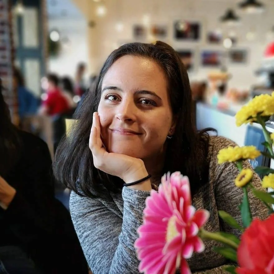 A woman with dark hair and a gray sweater sitting at a table with colorful flowers, smiling softly at the camera in a lively indoor setting.