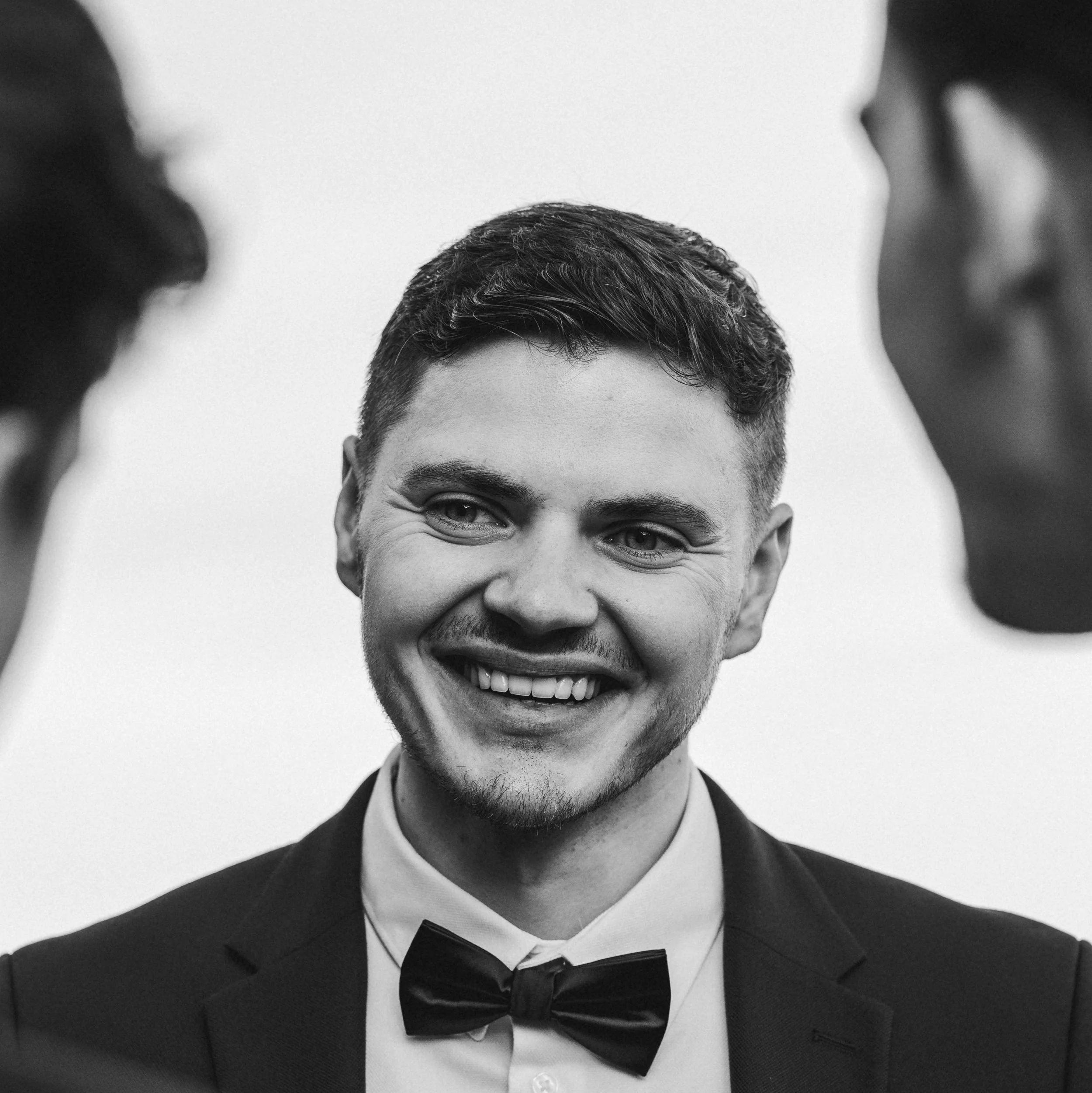 Un homme souriant portant un costume noir et un nœud papillon, vue partielle de la tête au-dessus des épaules.