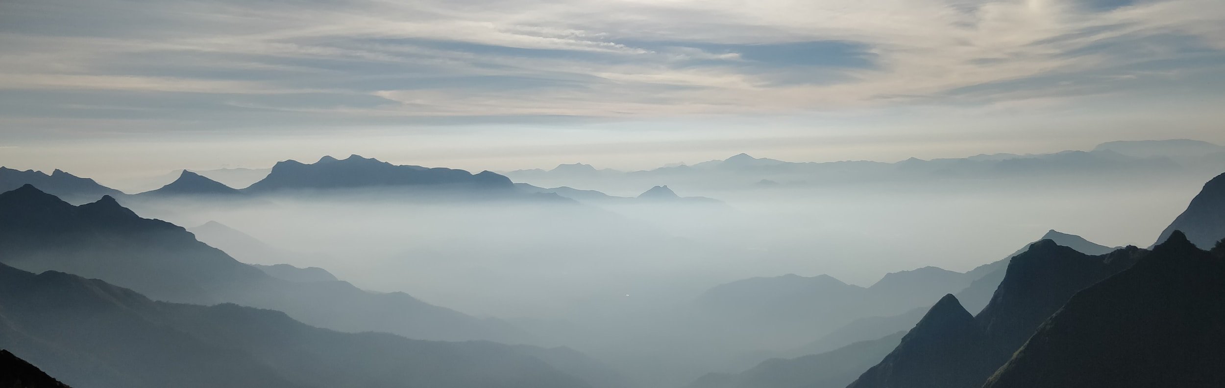Paysage de montagnes avec brume ou nuages bas, ciel partiellement nuageux, silhouettes de montagnes en arrière-plan.