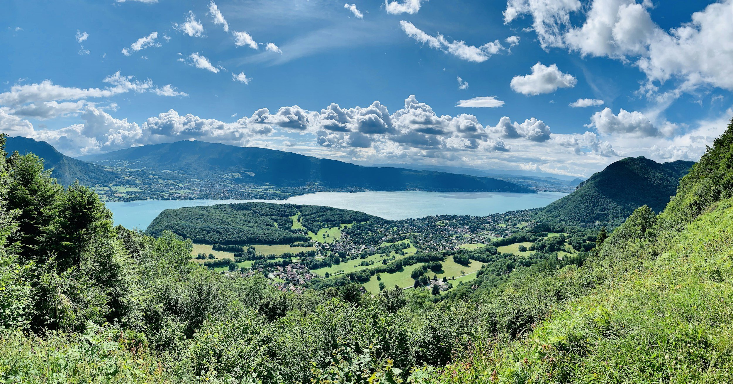 Paysage de montagnes avec un lac au centre, entouré de verdure et de nuages dans un ciel bleu.