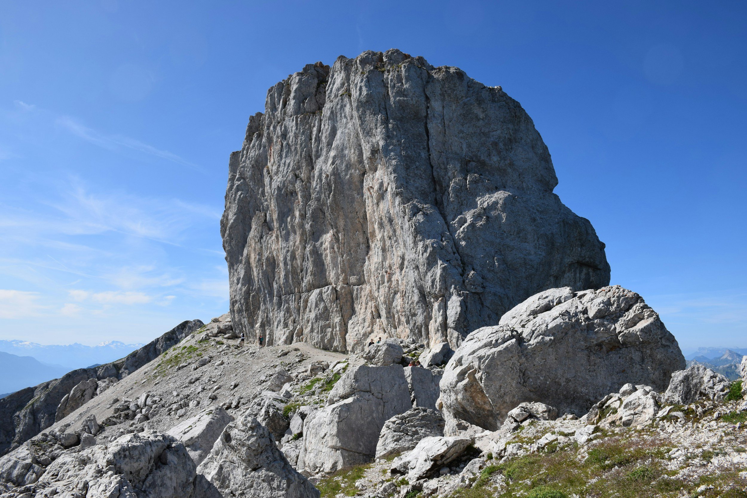 Une montagne rocheuse imposante sous un ciel bleu clair avec quelques nuages. La zone autour est rocheuse, avec de petites pierres et peu de végétation.