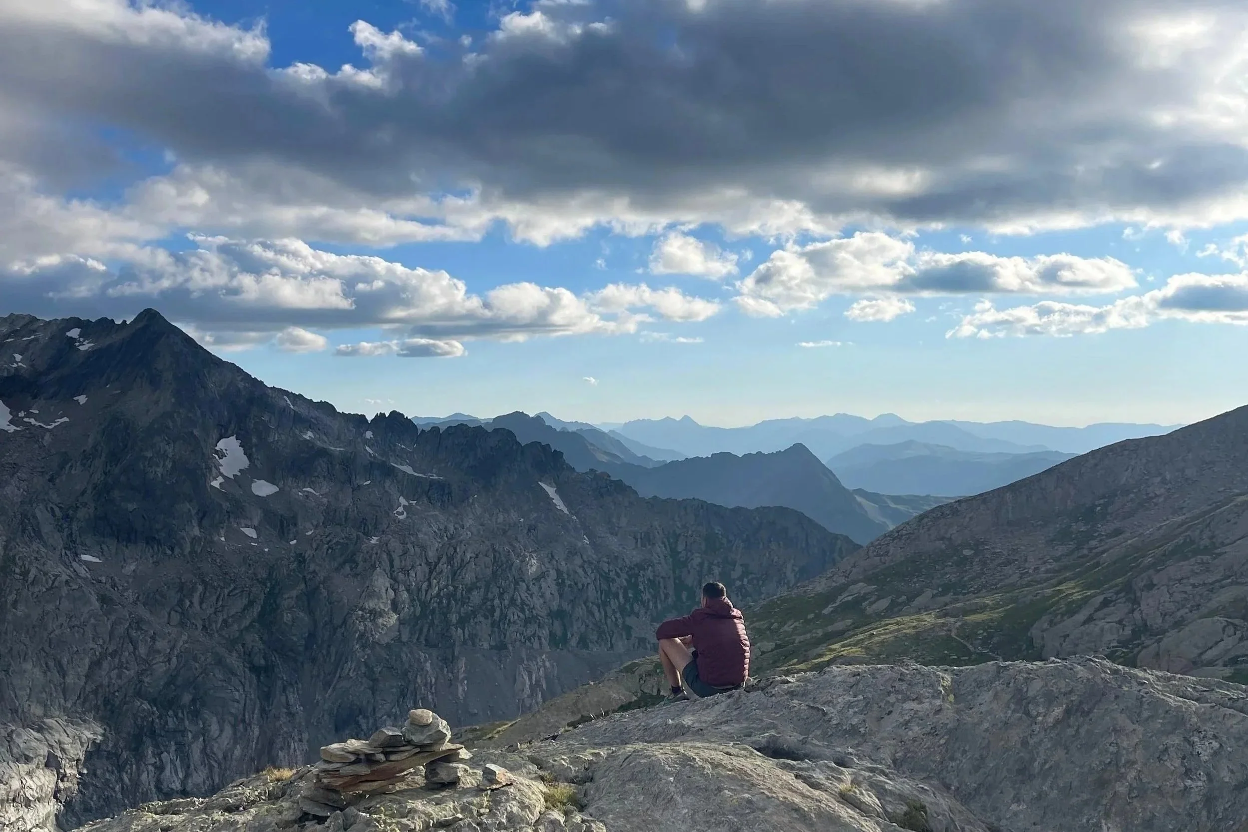 Une personne assise sur une roche regarde un paysage de montagnes rocheuses avec des sommets enneigés, sous un ciel partiellement nuageux.