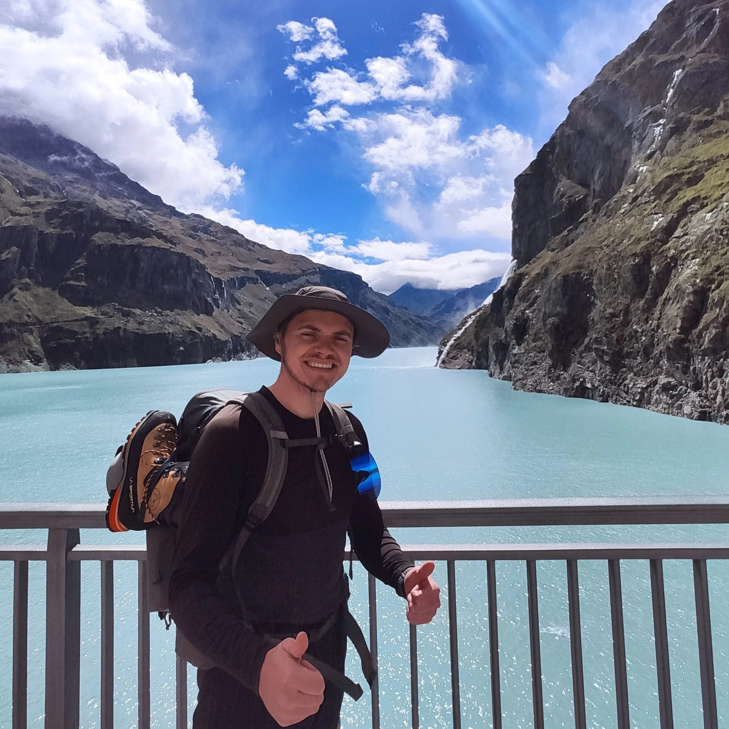 Un homme souriant avec un chapeau de randonnée pose devant un lac turquoise entouré de montagnes rocheuses et de nuages dans un paysage de montagne.