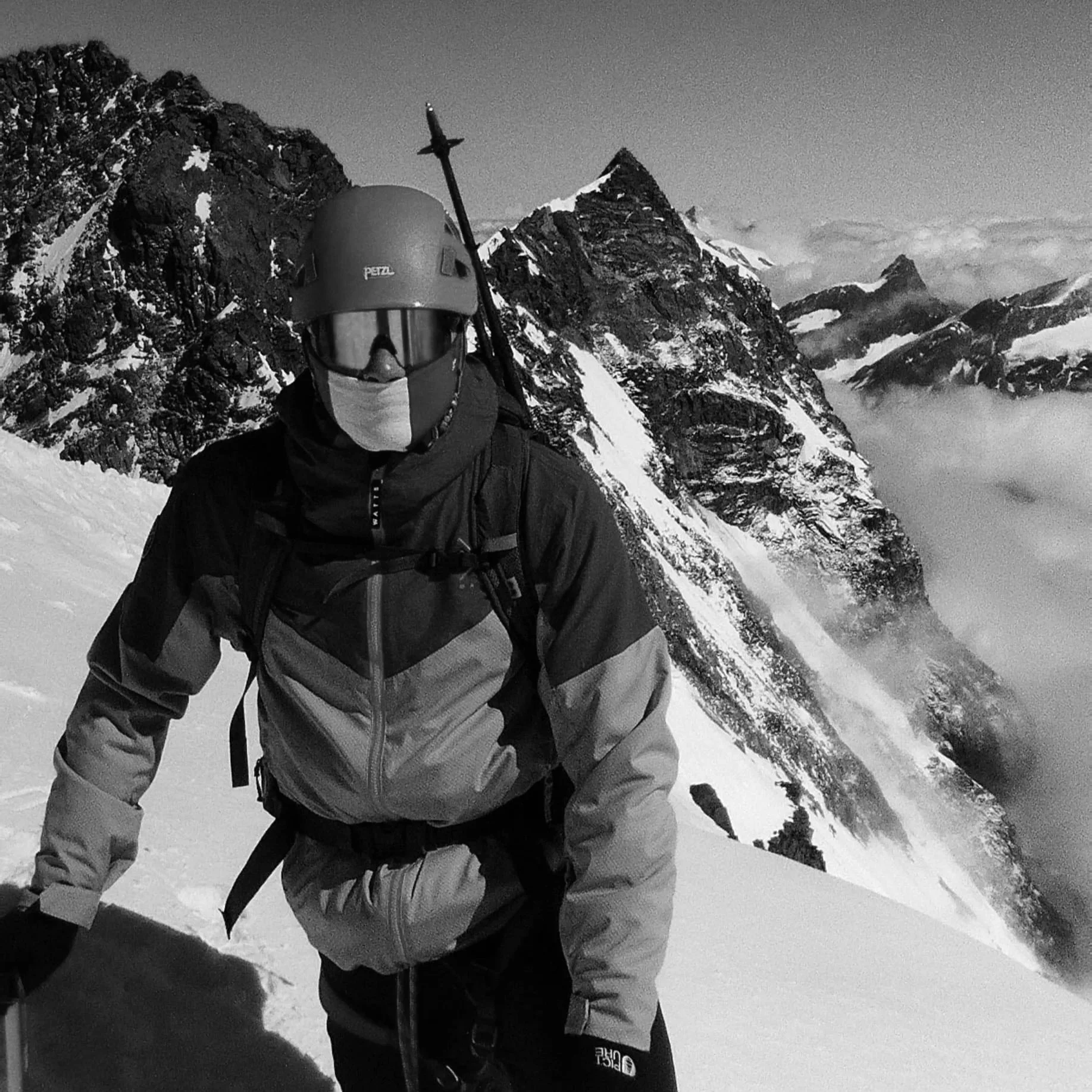 Un alpiniste portant un casque, masque et veste d'hiver, en cours d'ascension dans un paysage de montagnes enneigées avec des sommets pointus et des nuages autour.