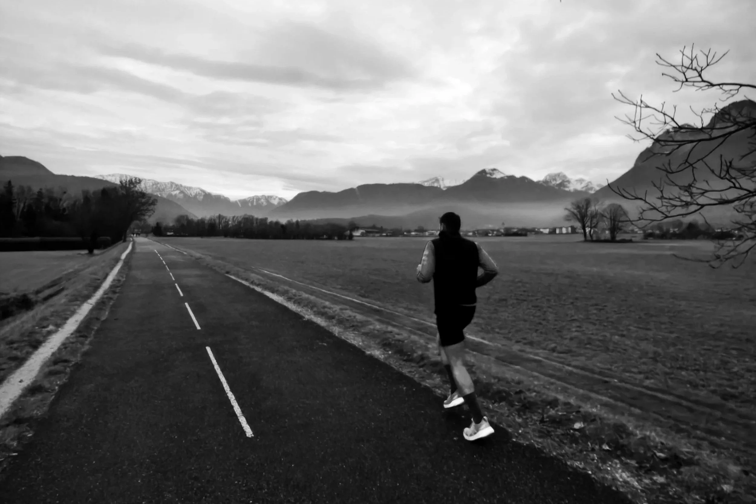 Un homme marche seul sur une route isolée dans un paysage montagneux, avec des montagnes enneigées au loin, sous un ciel nuageux.