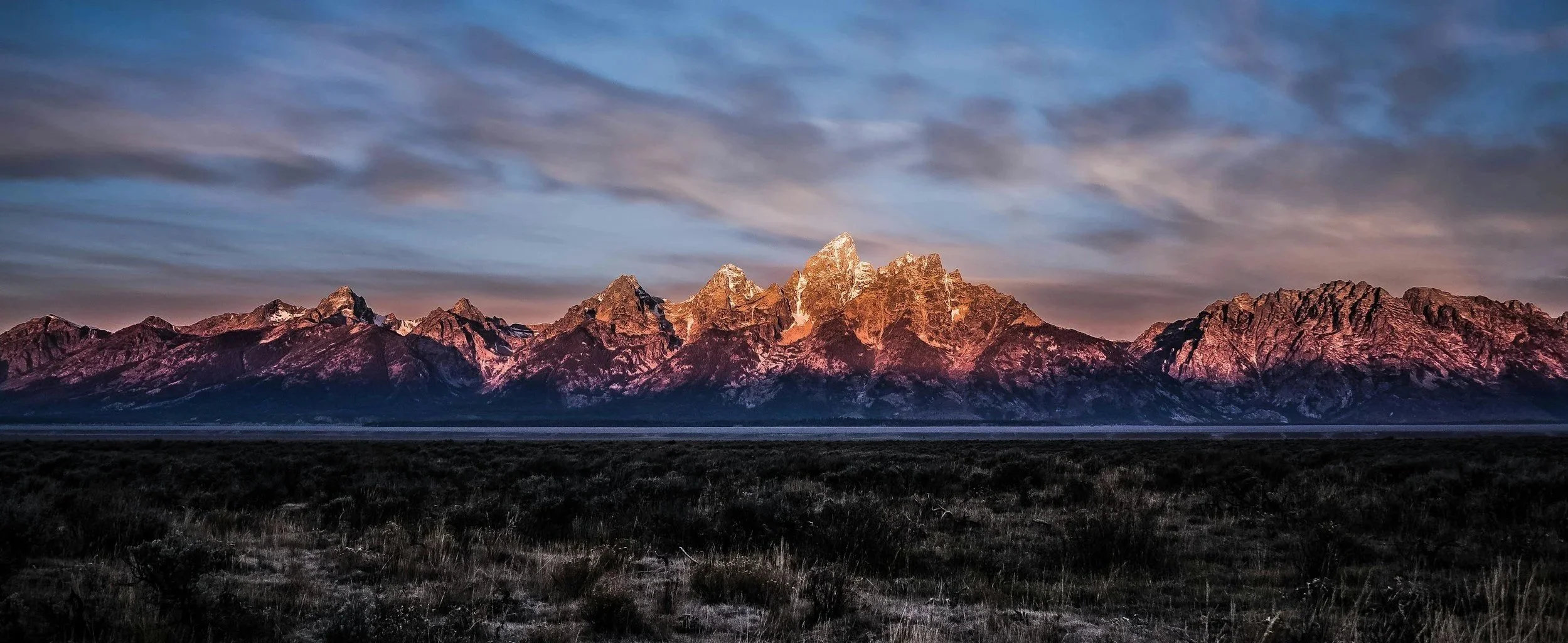 Vue panoramique de montagnes rocheuses au coucher du soleil, avec des nuages dans le ciel et un paysage de plaines au premier plan.