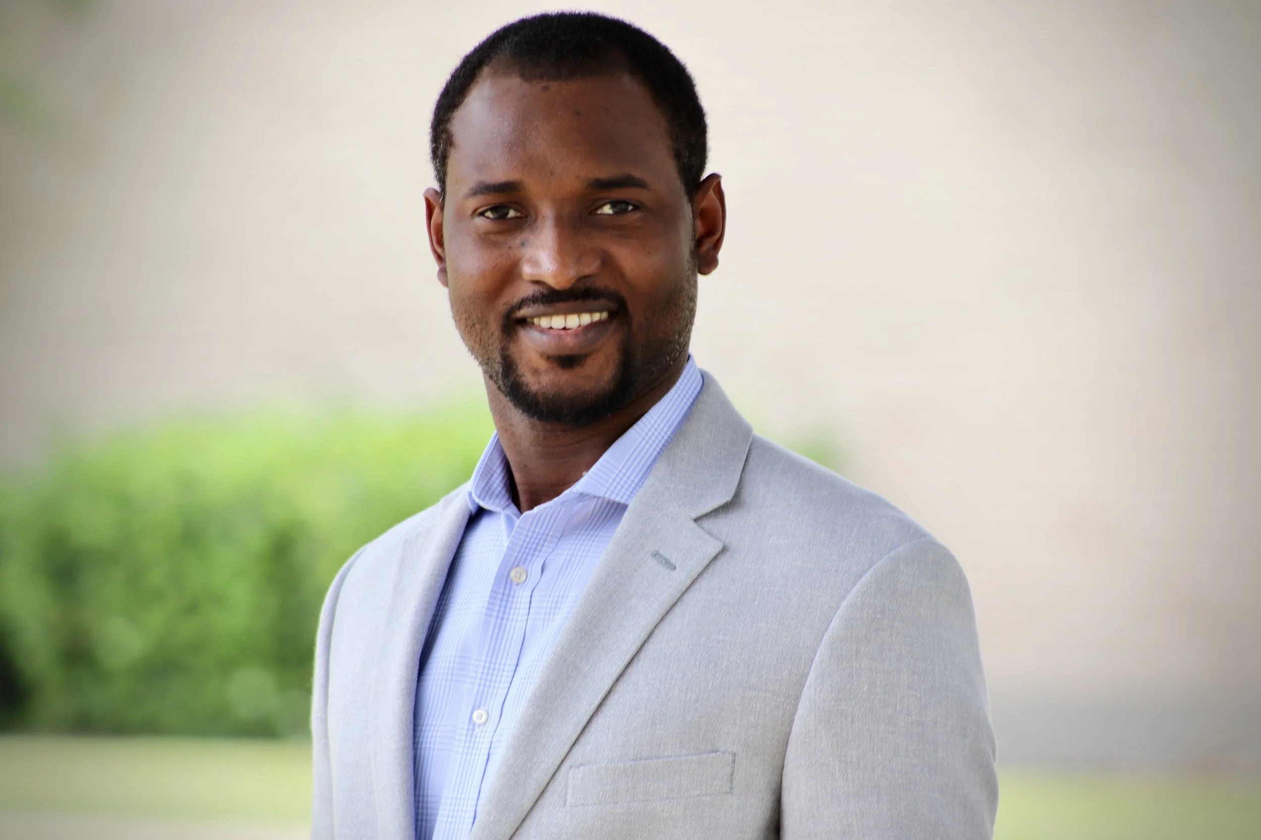A smiling man with dark skin and short hair wearing a light gray blazer and a collared shirt, standing outdoors with a blurred green background.