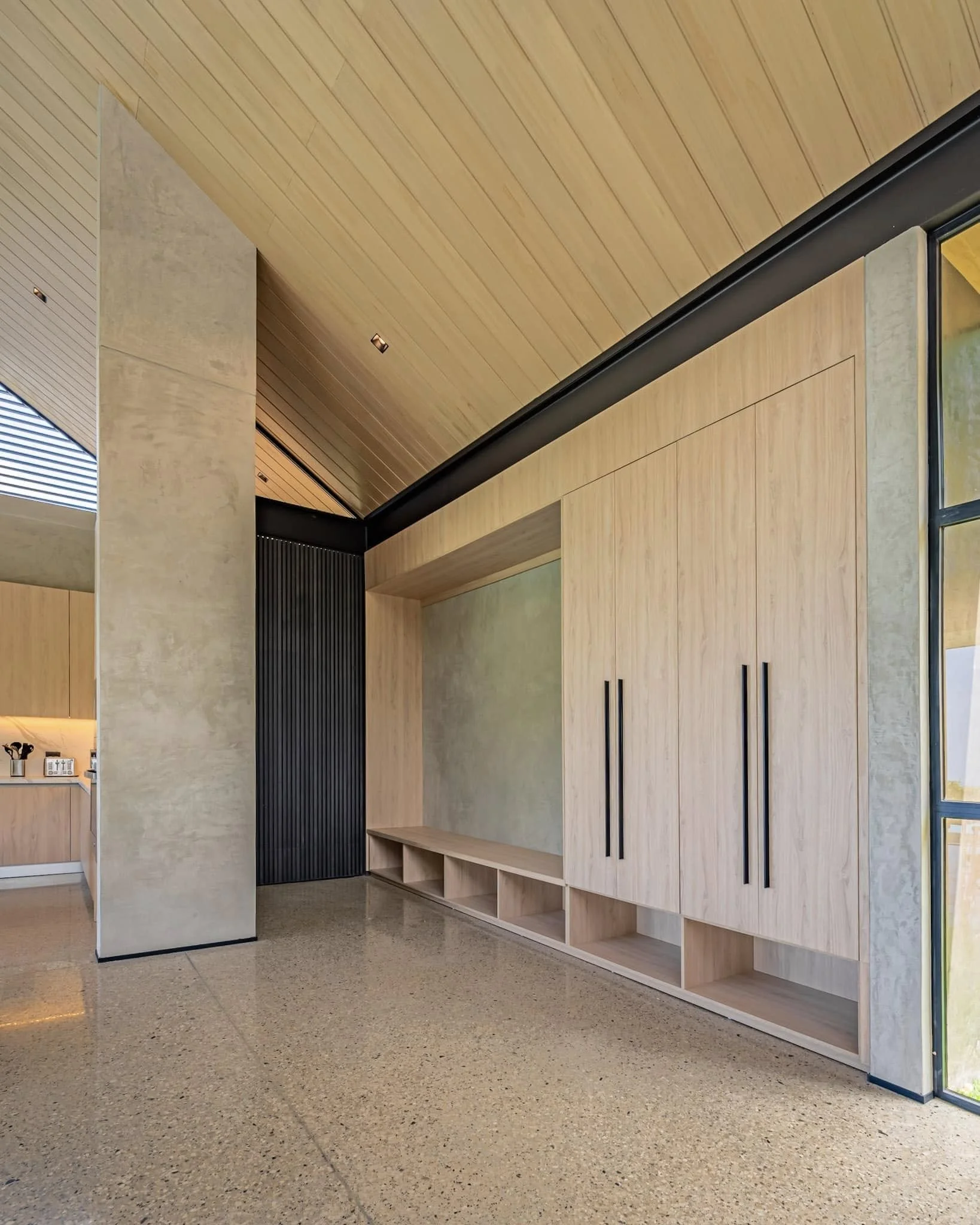 Interior space with wooden ceiling and walls, a tall light beige column, and built-in wooden storage with black handles, lighted by small ceiling lights.
