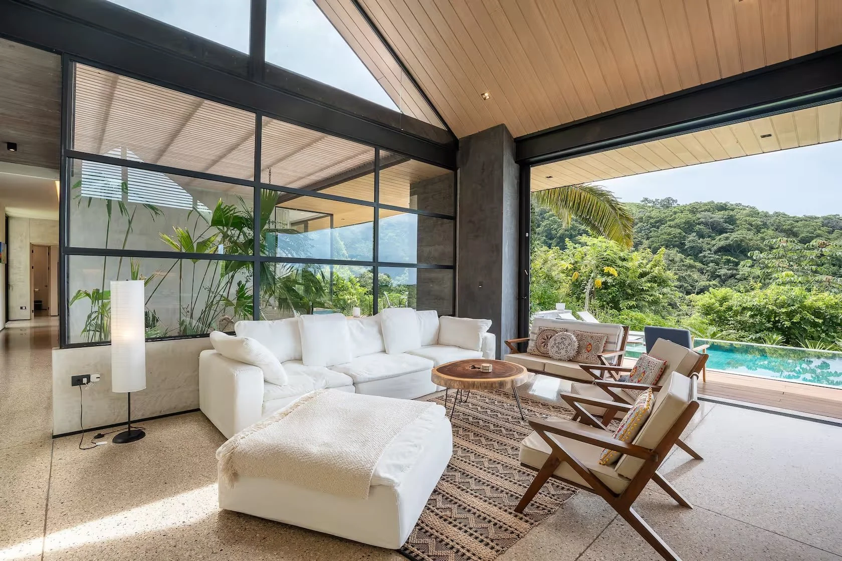 Modern living room with large glass windows, white sofa, wooden chairs, indoor plants, view of lush green trees, pool, and outdoor patio.