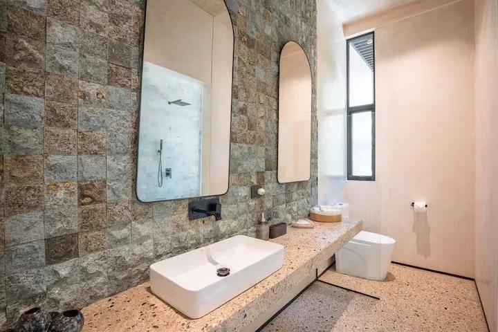 Modern bathroom with a stone accent wall, two tall mirrors, a rectangular vessel sink, a countertop, a toilet, and a large window