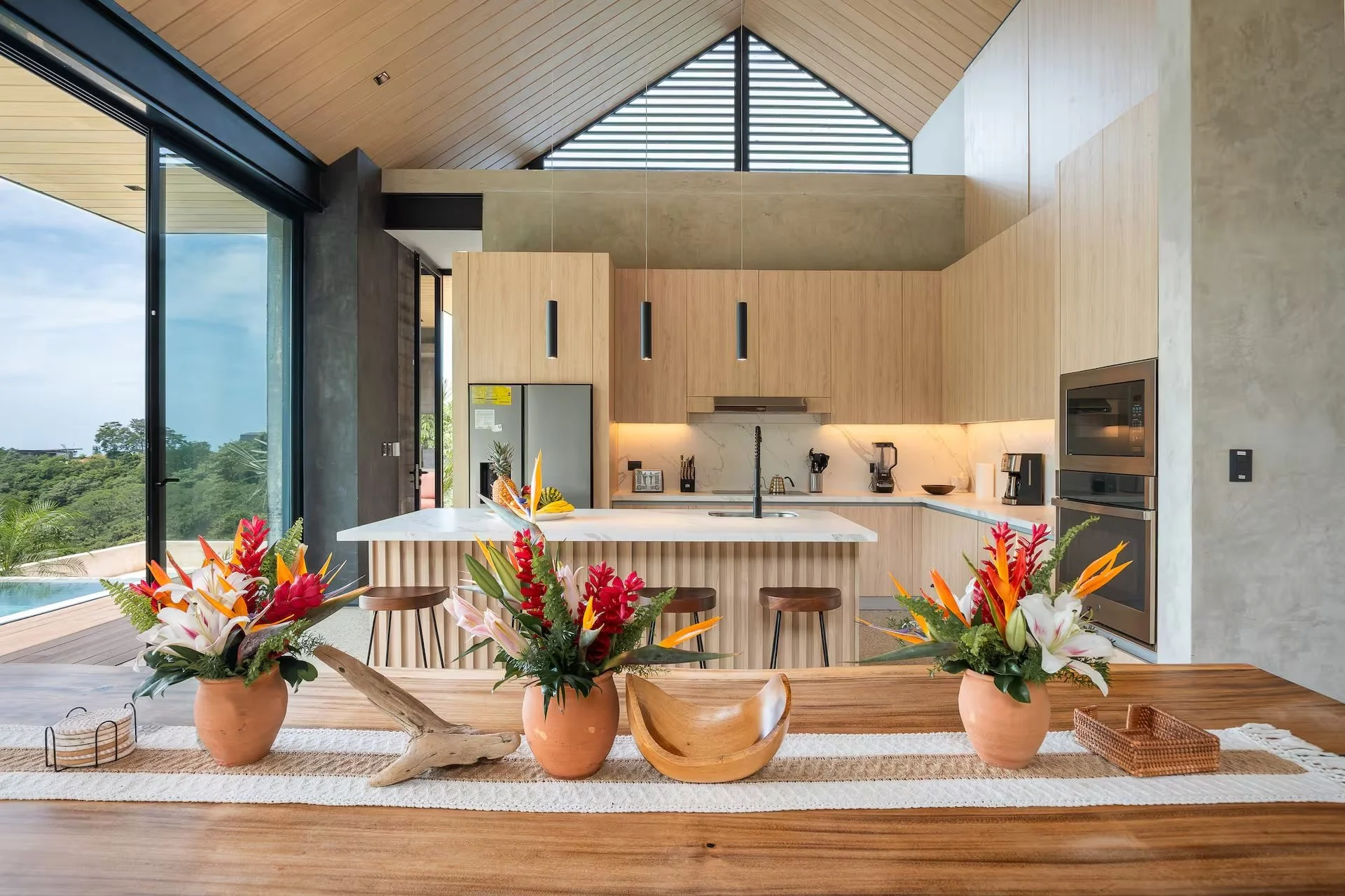 Modern kitchen with wooden cabinets and a white marble island, decorated with colorful tropical flower arrangements in terracotta pots, overlooking an outdoor swimming pool and greenery.