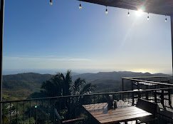 Outdoor patio with wooden table and chairs overlooking lush green hills and a body of water in the distance under a clear blue sky.