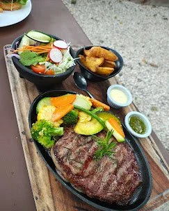 A meal featuring a grilled steak topped with herbs, accompanied by a side salad and a small bowl of crispy fried chicken tenders with dipping sauces on a wooden table.