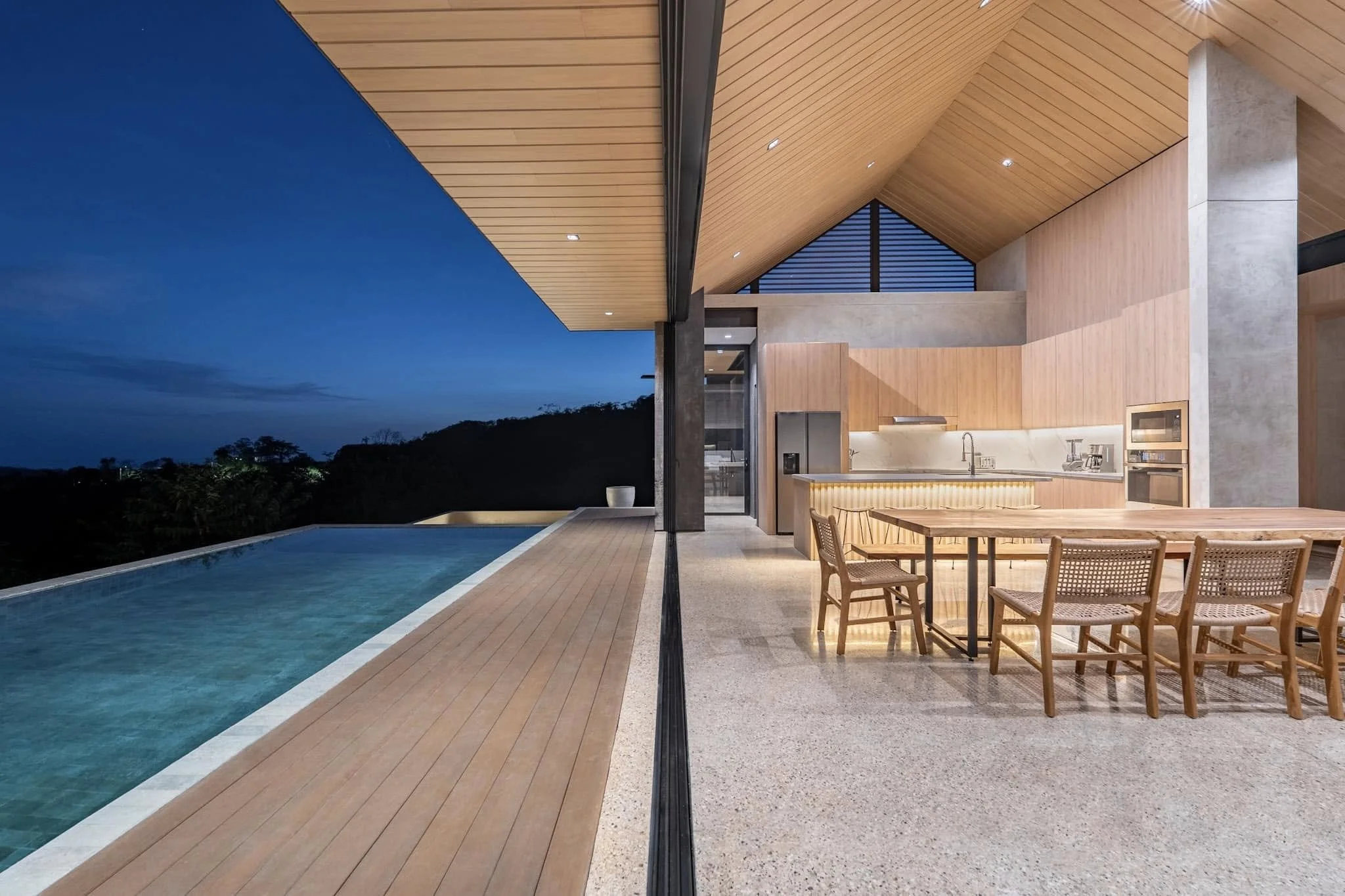 Modern house with wooden and concrete exterior, a large open kitchen, dining area, and an outdoor pool at night with landscape and trees in the background.