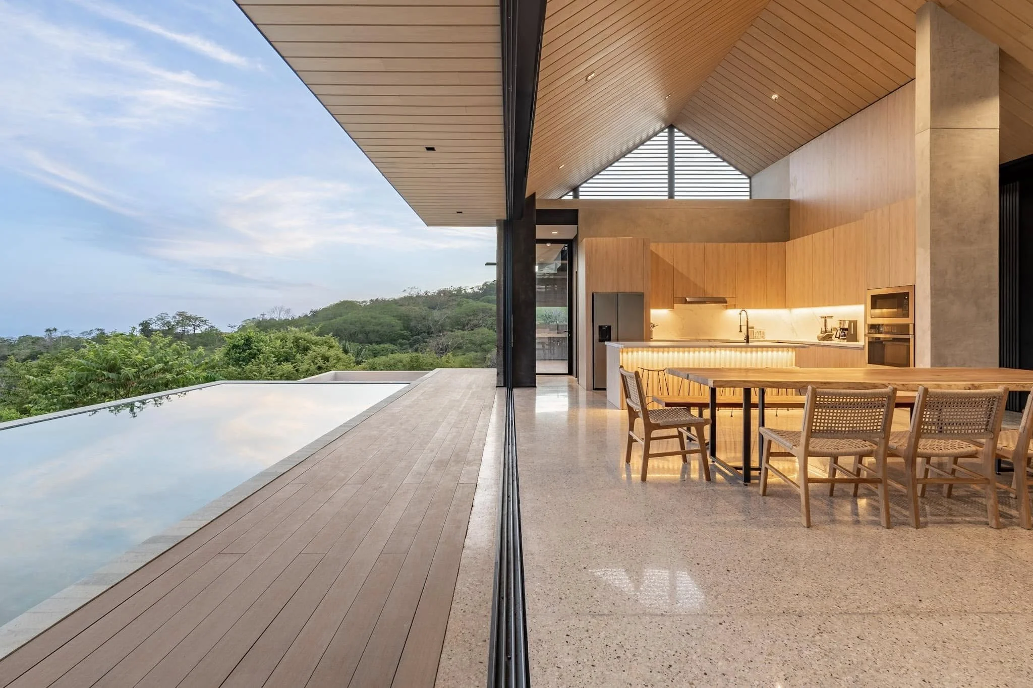 Modern indoor living space with wood-paneled ceiling and large windows showing a forest landscape, with a dining area and an infinity pool on a wooden deck.