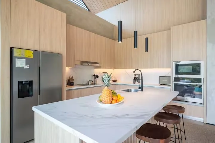 Modern kitchen with light wood cabinets, a white marble island with seating, stainless steel refrigerator, built-in oven, and microwave, decorative fruit bowl with pineapple and oranges, hanging pendant lights, and a minimalistic design.