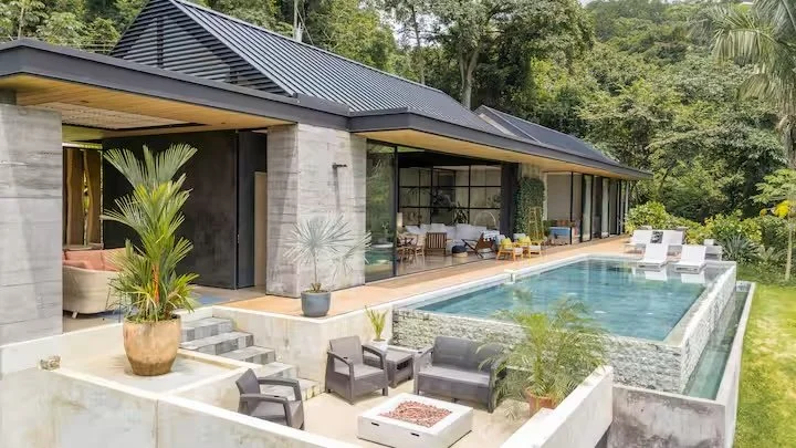 Modern house with large glass windows and a swimming pool in the backyard, surrounded by greenery and outdoor furniture.