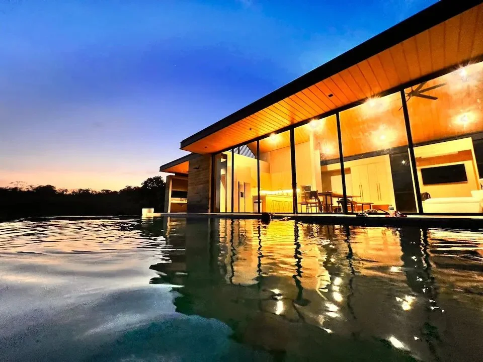 Modern house with large glass windows and interior lighting, situated next to a swimming pool at dusk, reflecting the house and the evening sky.