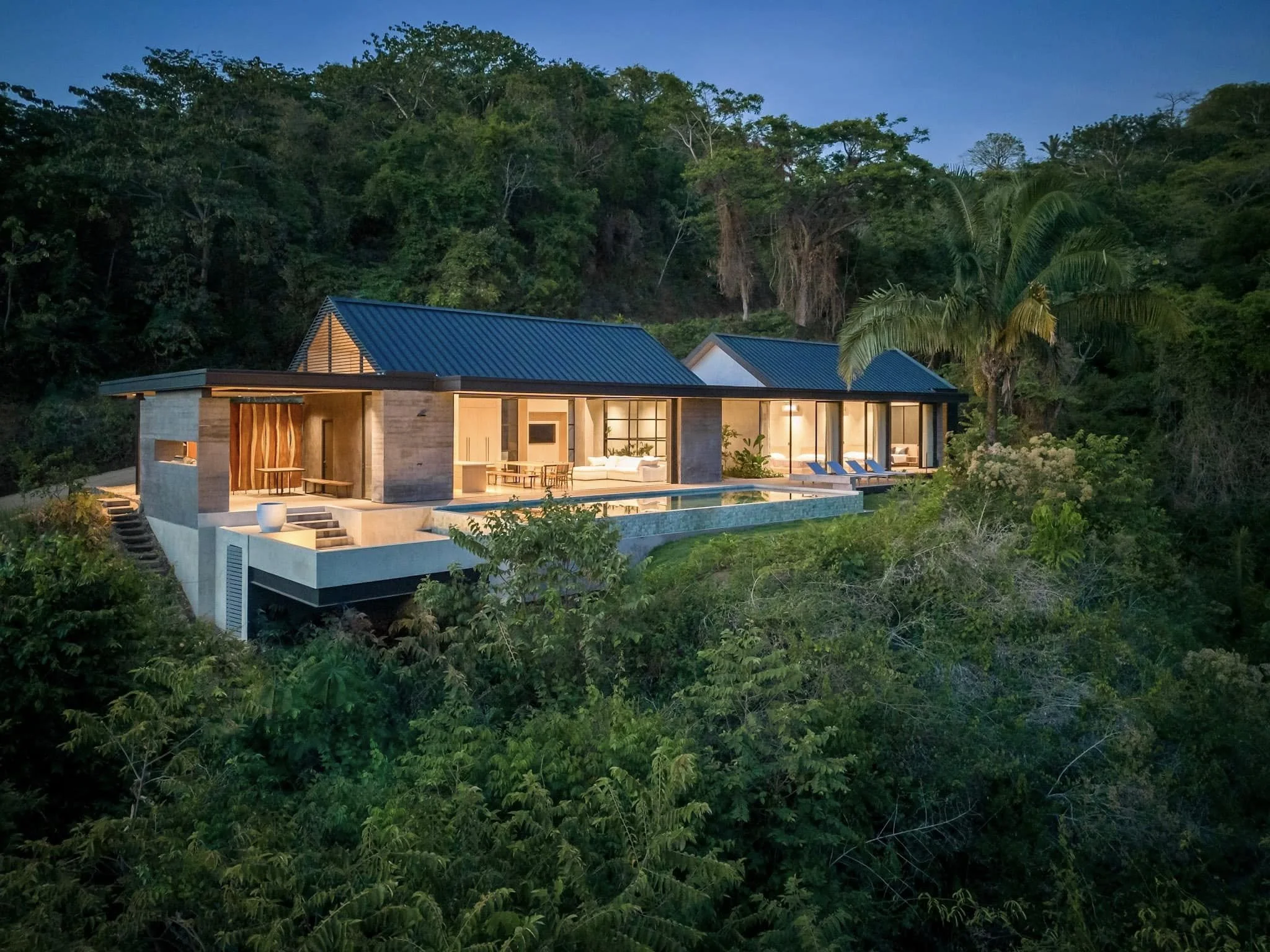 Modern house with large windows and a swimming pool, surrounded by lush green trees and plants, at dusk.