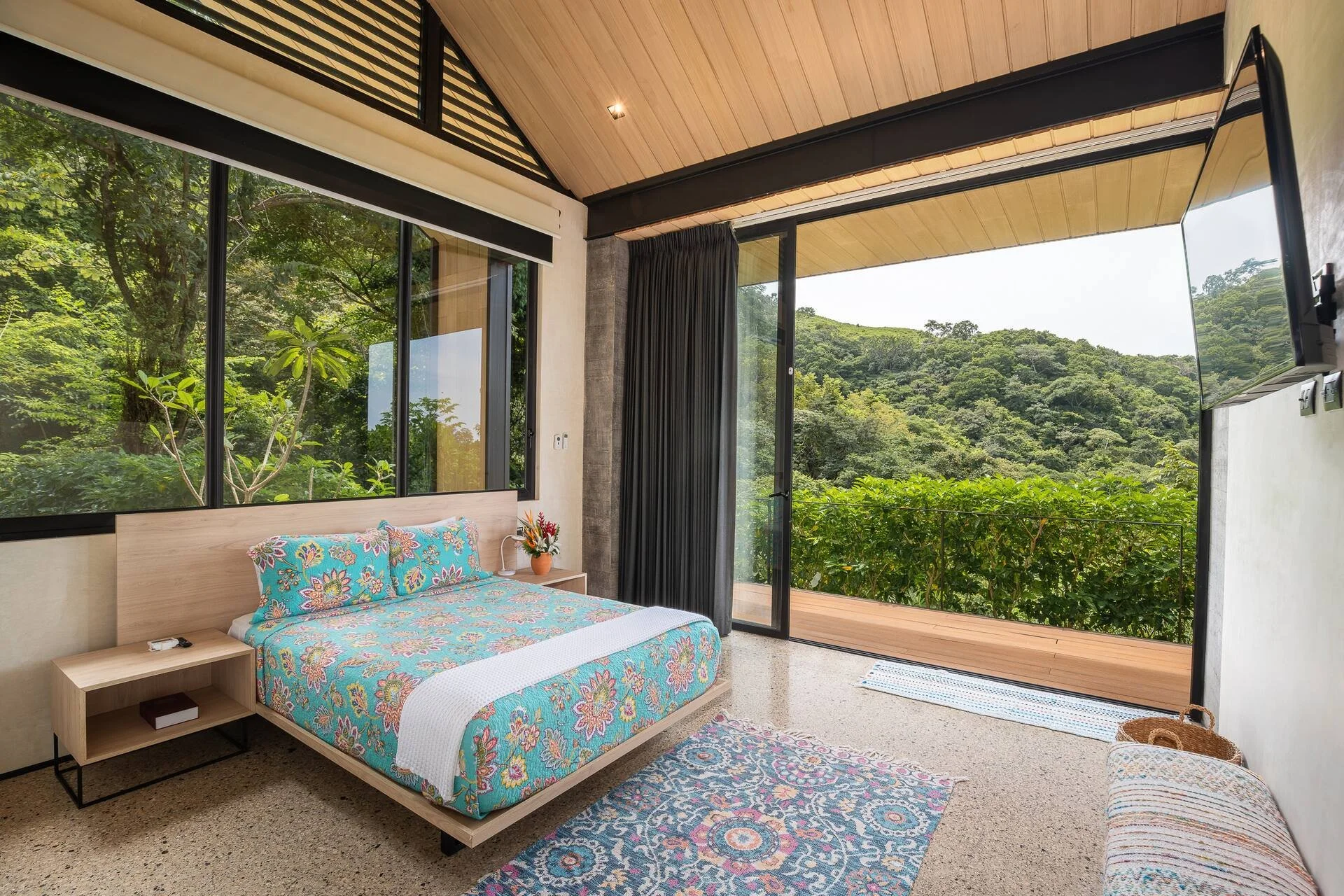 A bedroom with large glass sliding doors, a bed with a colorful floral quilt, a bedside table with books and a plant, a patterned rug, and a view of green hills and trees outside.