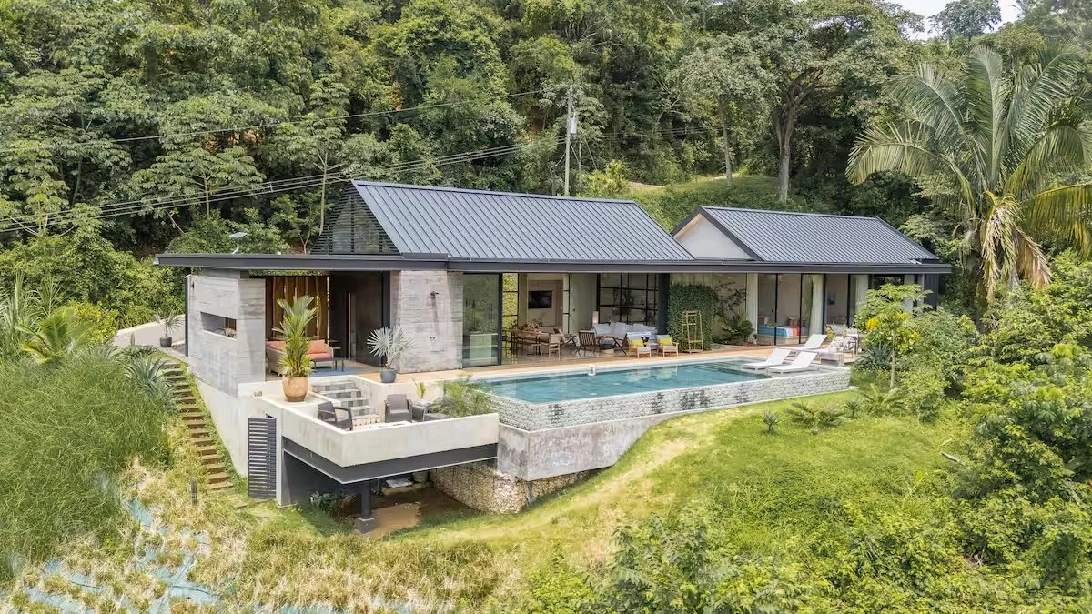 A modern house with a dark metal roof, large glass windows, and an outdoor pool, surrounded by lush green trees on a hillside.