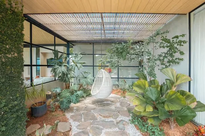 Indoor patio with large leafy plants and a hanging egg-shaped chair in the center, surrounded by stone pavers and glass walls.
