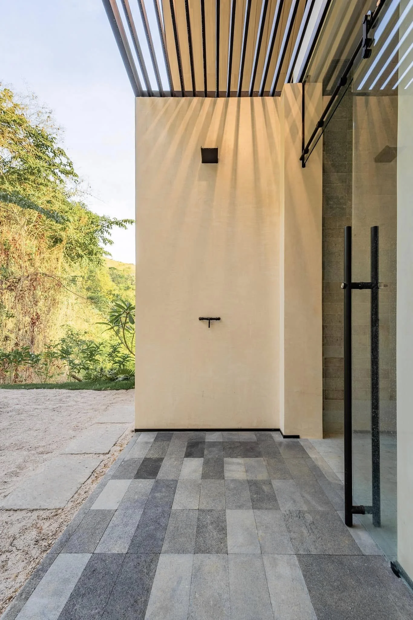 Modern building exterior with a beige wall, glass door, black metal railing, and outdoor tiled patio under a striped roof, with trees and greenery in the background.