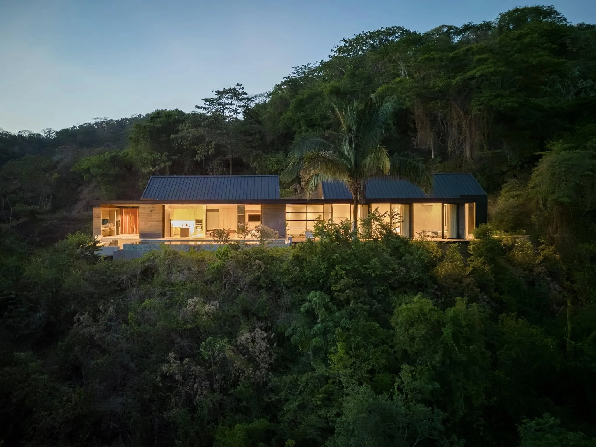 A modern house with large windows and a metal roof is illuminated from inside, set against a lush green hillside with trees.