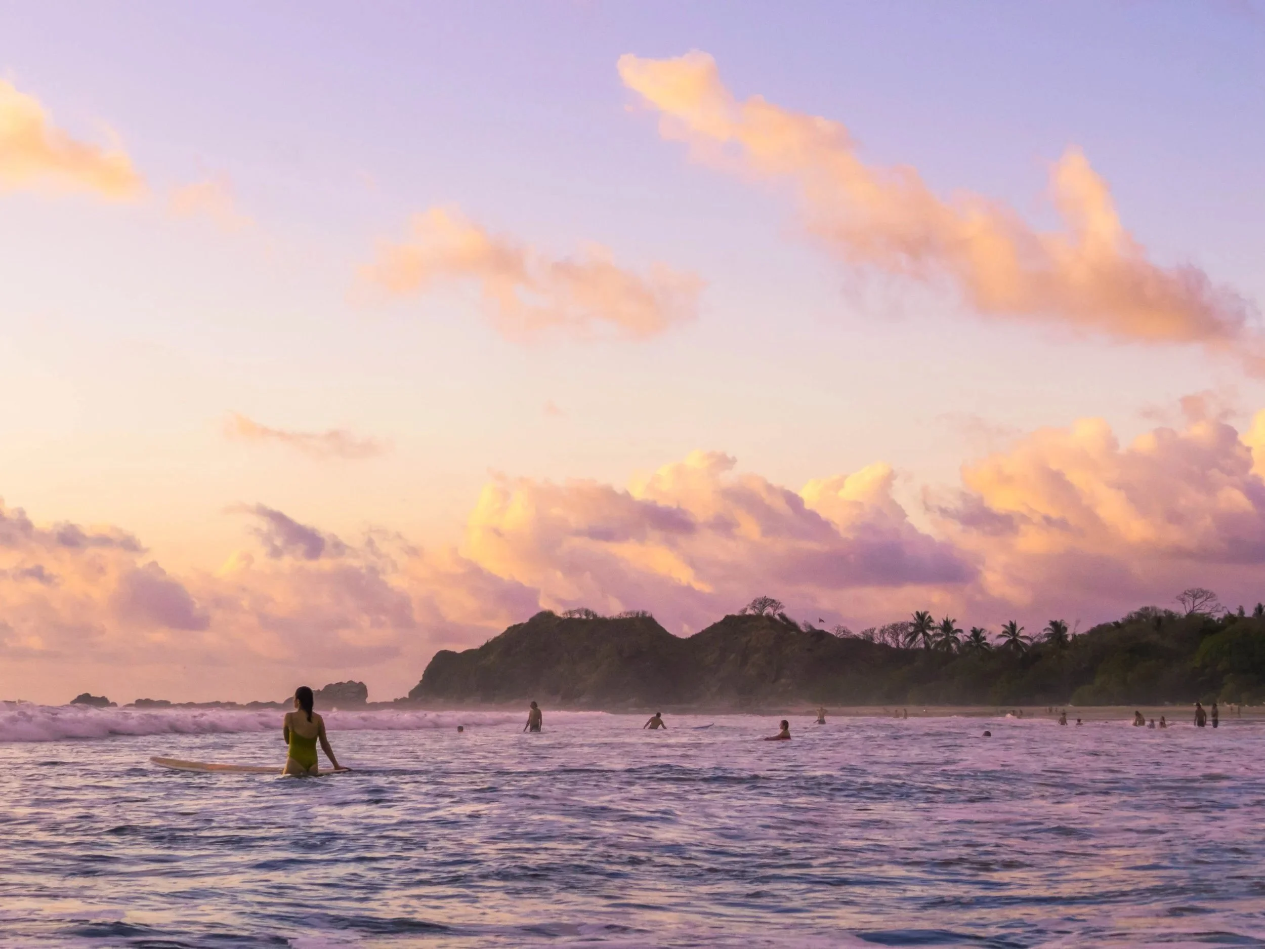 People enjoying the ocean at sunset with a mountain in the background, pink and purple sky, and calm water.