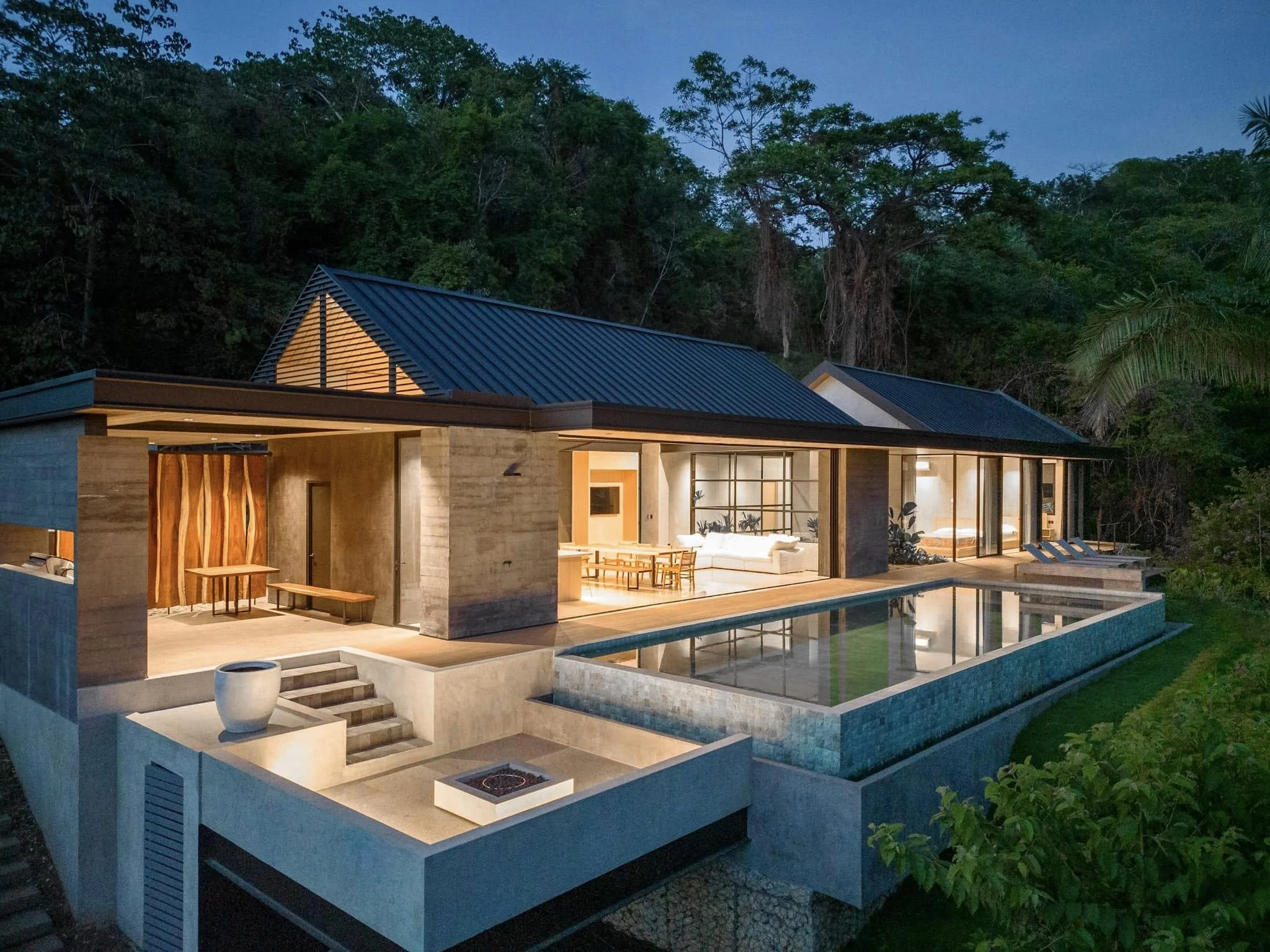 Modern house with a blue metal roof and large glass windows, surrounded by lush green trees, during twilight, with outdoor seating and a pool.