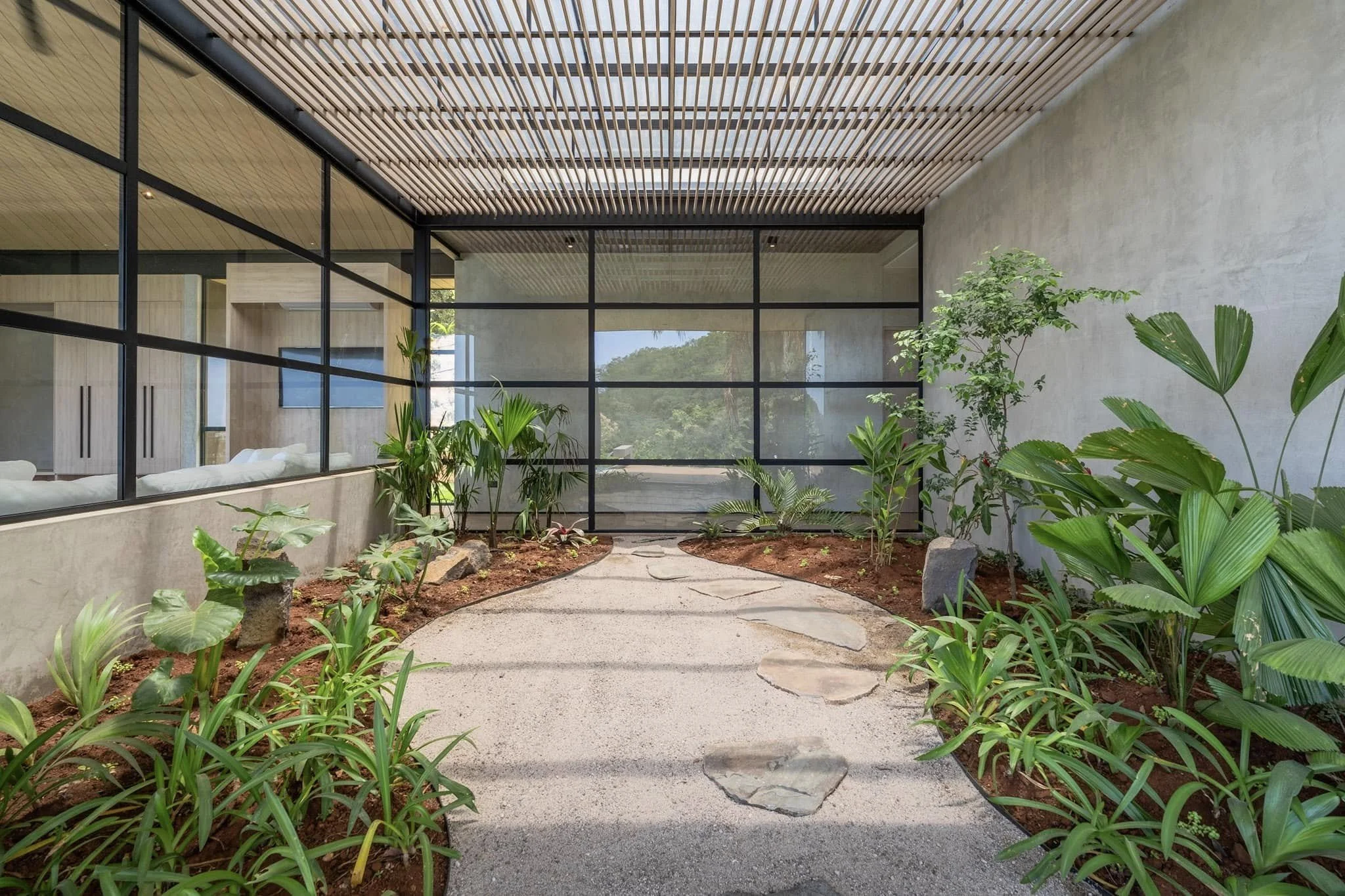 Indoor garden with a concrete pathway surrounded by various green plants and trees, enclosed by glass walls and a ceiling with wooden slats.