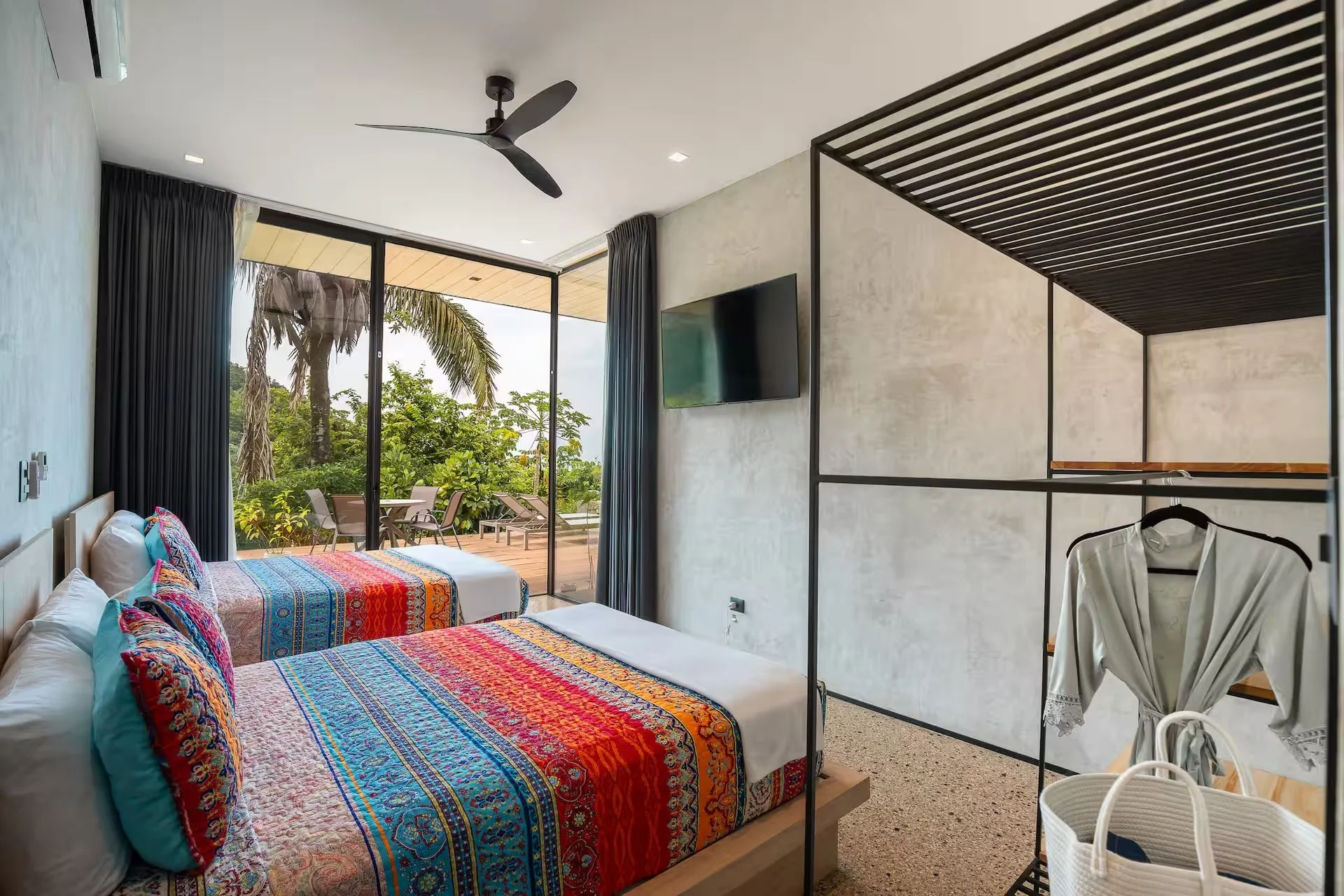 A modern hotel room with two beds covered with colorful, patterned bedspreads. Floor to ceiling glass doors open to an outdoor balcony with lounge chairs and greenery, including palm trees. A black ceiling fan hangs from the white ceiling. A flat-screen TV is mounted on the wall, and a black metal frame divider separates part of the room. There is a white robe hanging on a rack and a white basket in the foreground.