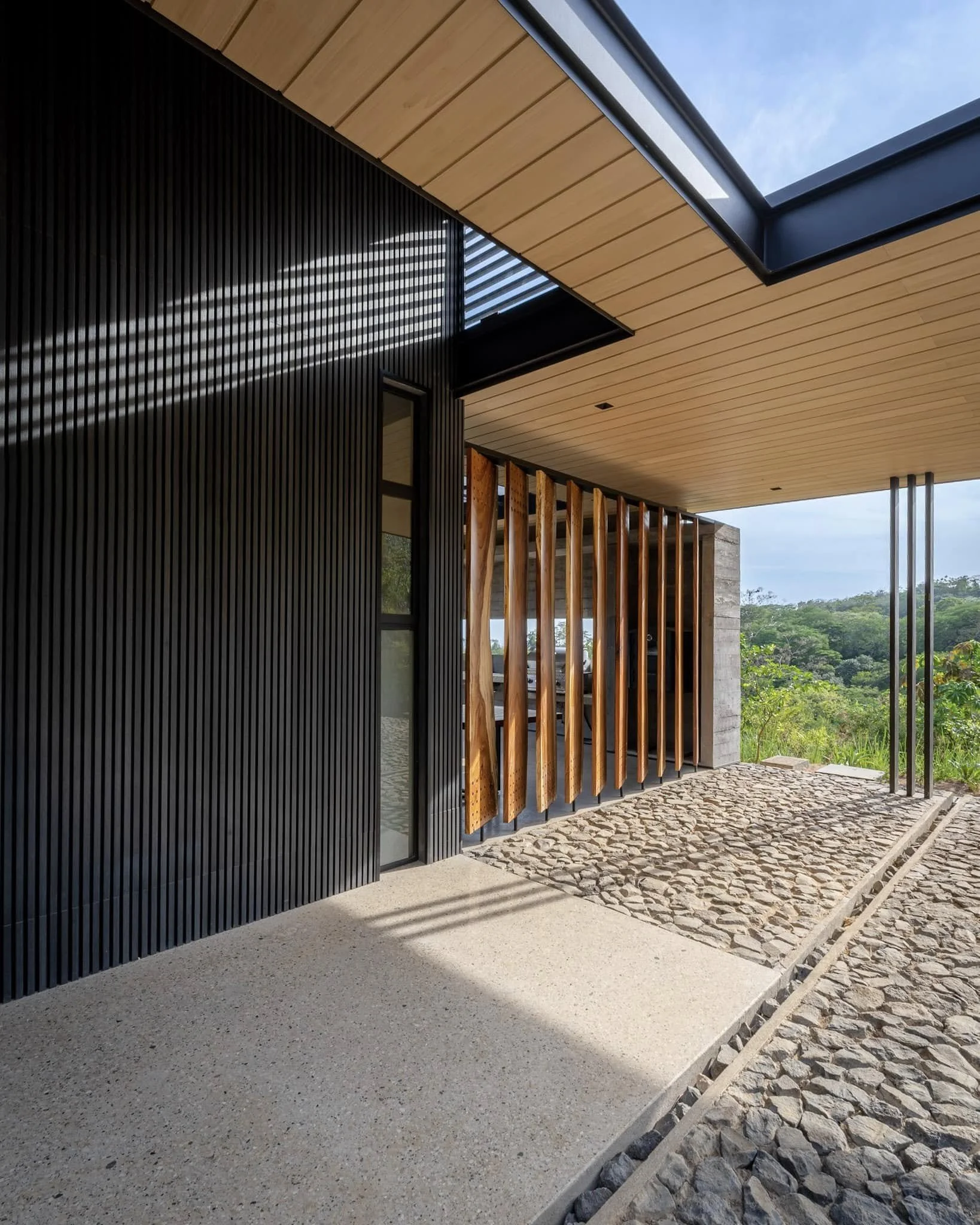 Modern building with black, wood, and stone exterior, large glass window, and a skylight in the ceiling. Outdoor area with cobblestone paving and a view of greenery and trees.