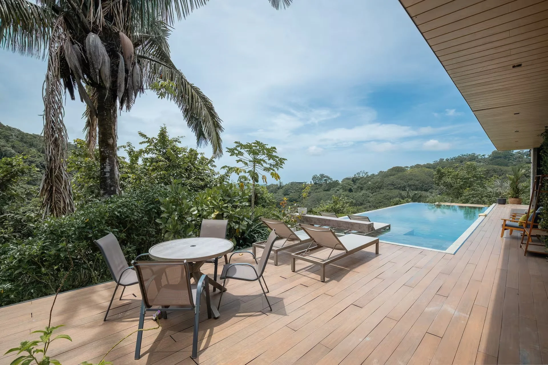 A modern outdoor patio featuring a rectangular swimming pool with a view of lush greenery and hills in the background. The patio has wooden flooring, lounge chairs, a round table with chairs, potted plants, and is partially shaded by a roof.