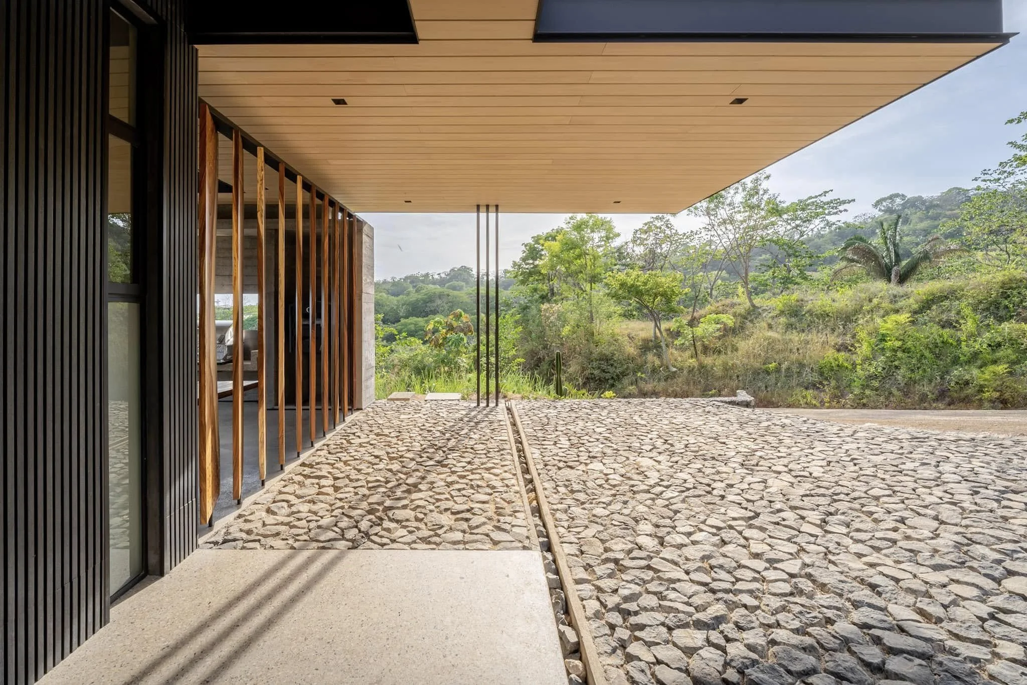 Modern building exterior with wooden slat accents and cobblestone flooring, overlooking lush green landscape and trees.